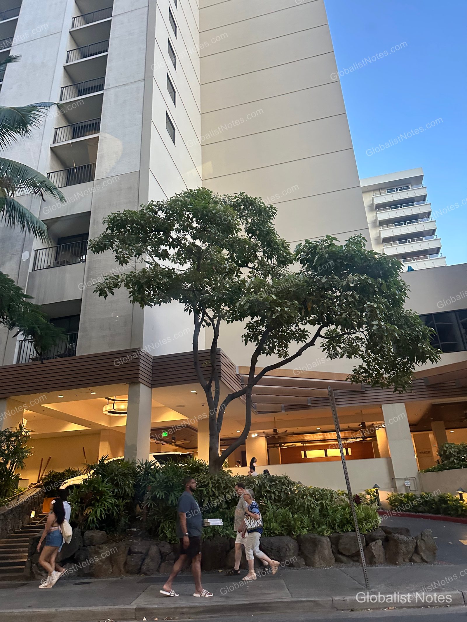 Exterior view of Hyatt Place Waikiki Beach in Honolulu, Hawaii, showing the building facade, landscaping at the entrance, and guests walking on the sidewalk.