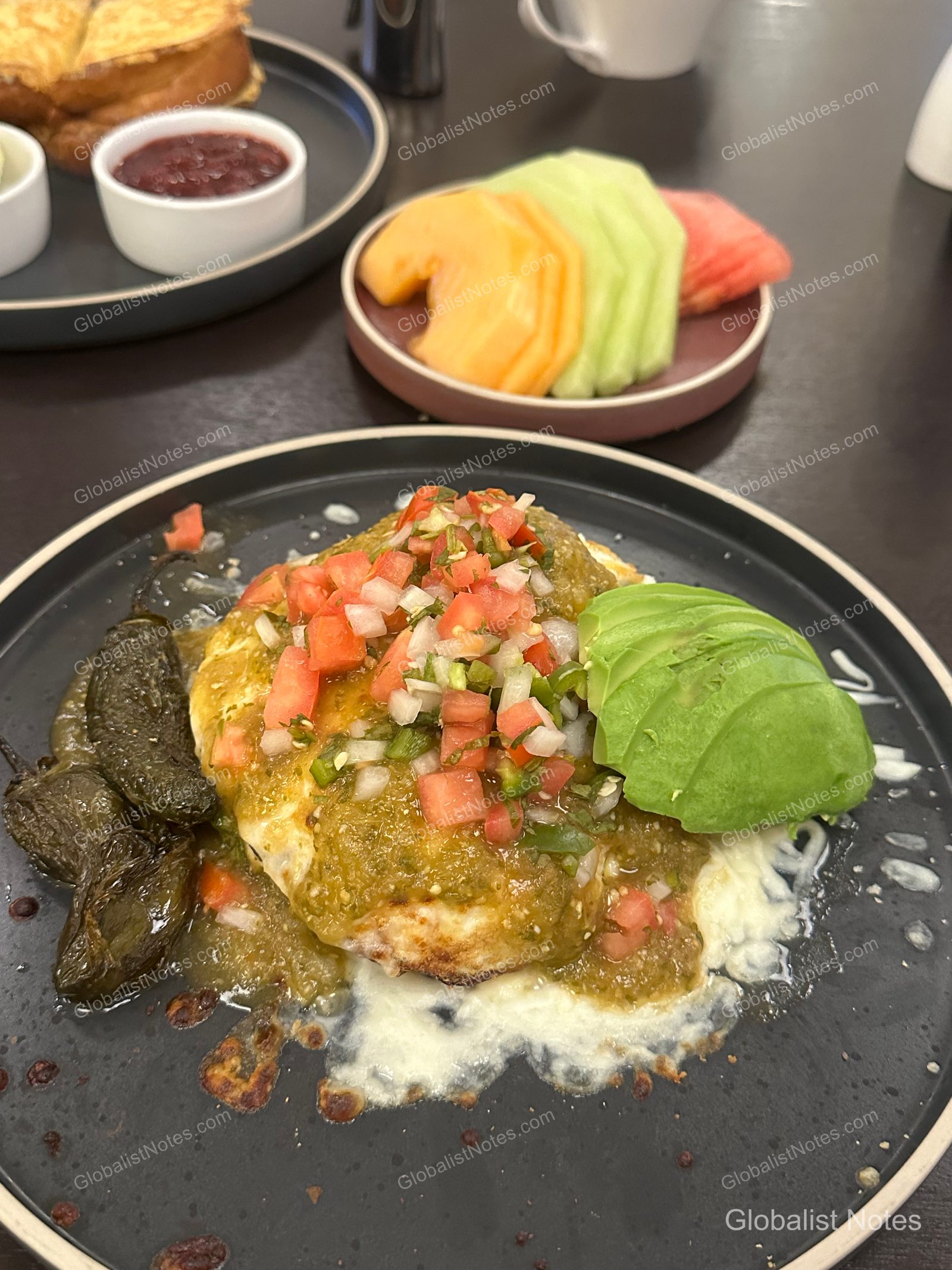 Breakfast at Hyatt Regency Los Angeles International Airport featuring an egg dish with salsa verde, tomato salsa, sliced avocado, and roasted jalapeño served on a black plate, with a side of fresh fruit and French toast in the background.