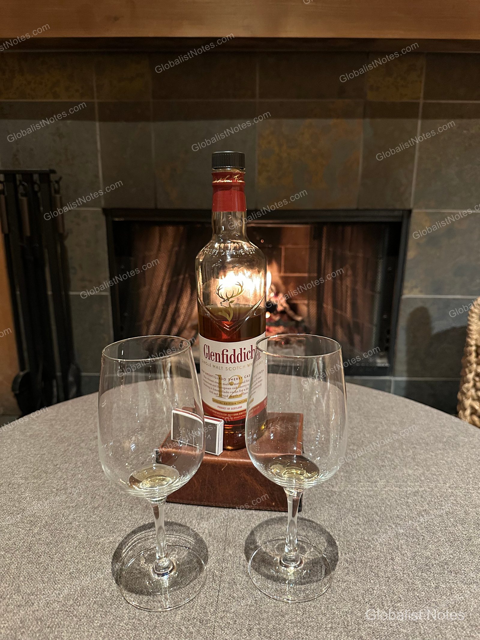 A bottle of Glenfiddich whisky and two empty glasses on a table in front of a fireplace in an Alila Ventana Big Sur Junior Suite.