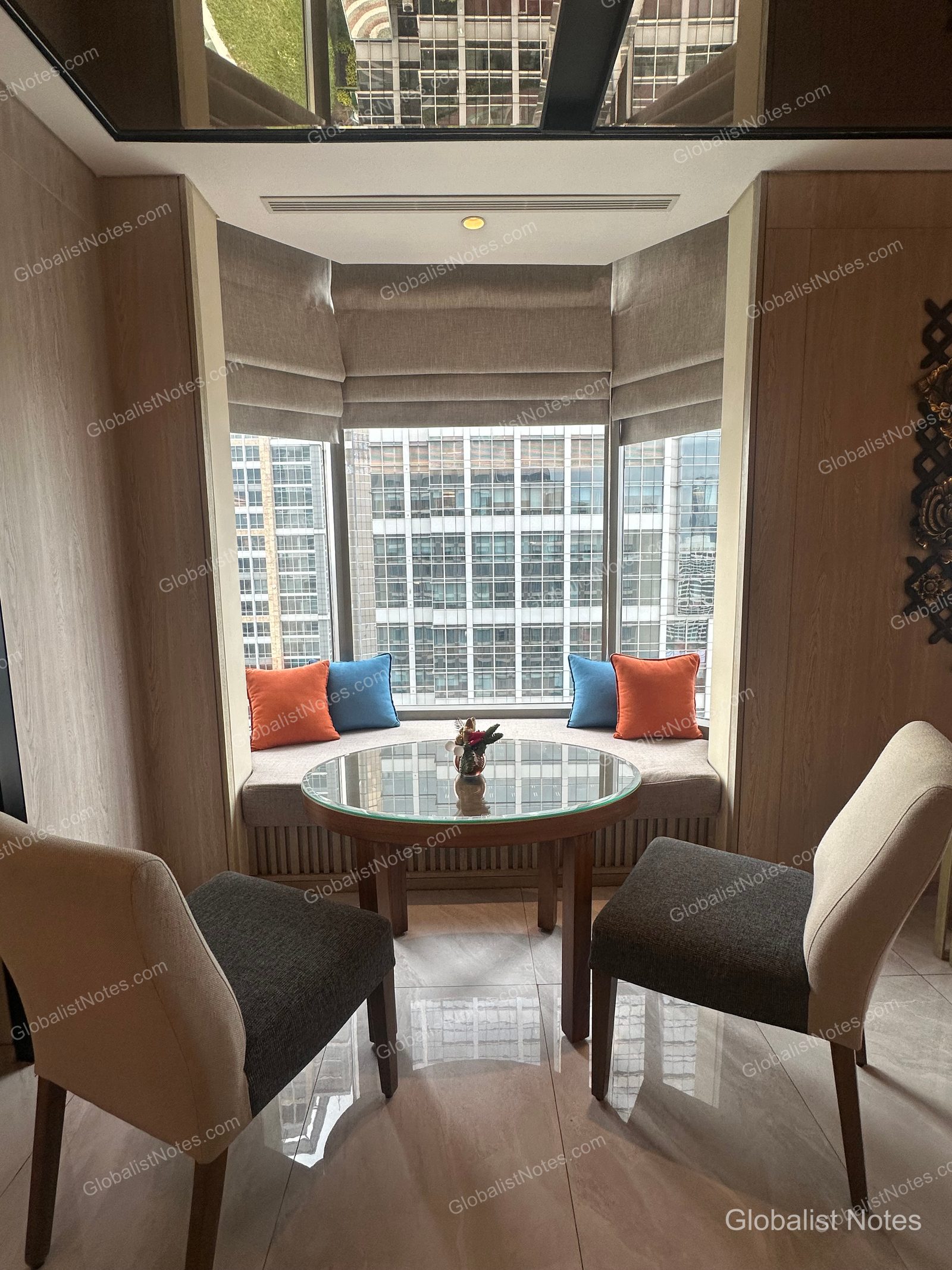 A cozy seating area by the window in a Grand Hyatt Jakarta guest room, featuring a long bench seat along the bay window, a round glass-topped table, and two chairs, with a view of Jakarta's high-rise buildings outside.