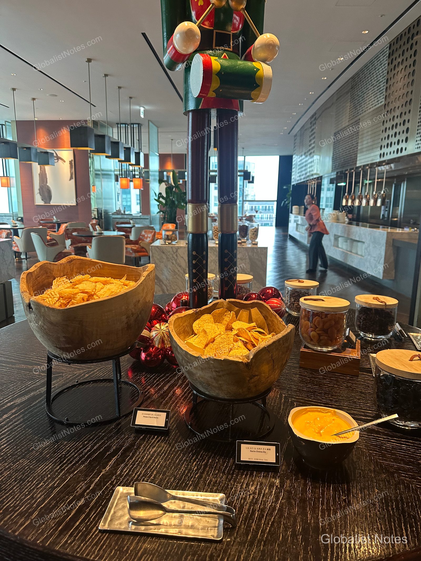 Nachos and potato chips in a wooden bowl on a table at the Grand Hyatt Jeju lounge, with a large nutcracker decoration and glass jars filled with various snacks in the background, along with lounge seating and lighting.