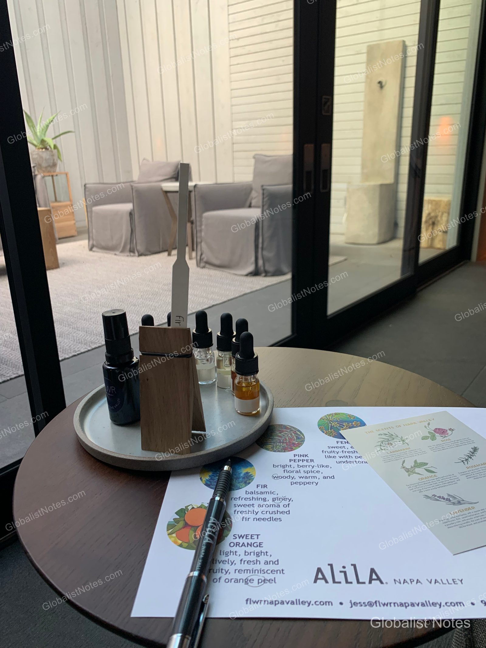A fragrance sampling kit and information card placed on a round table inside an Alila Napa Valley guest room. In the background, floor-to-ceiling windows look out onto an outdoor terrace with armchairs and sculptures, showcasing the serene Napa Valley landscape.