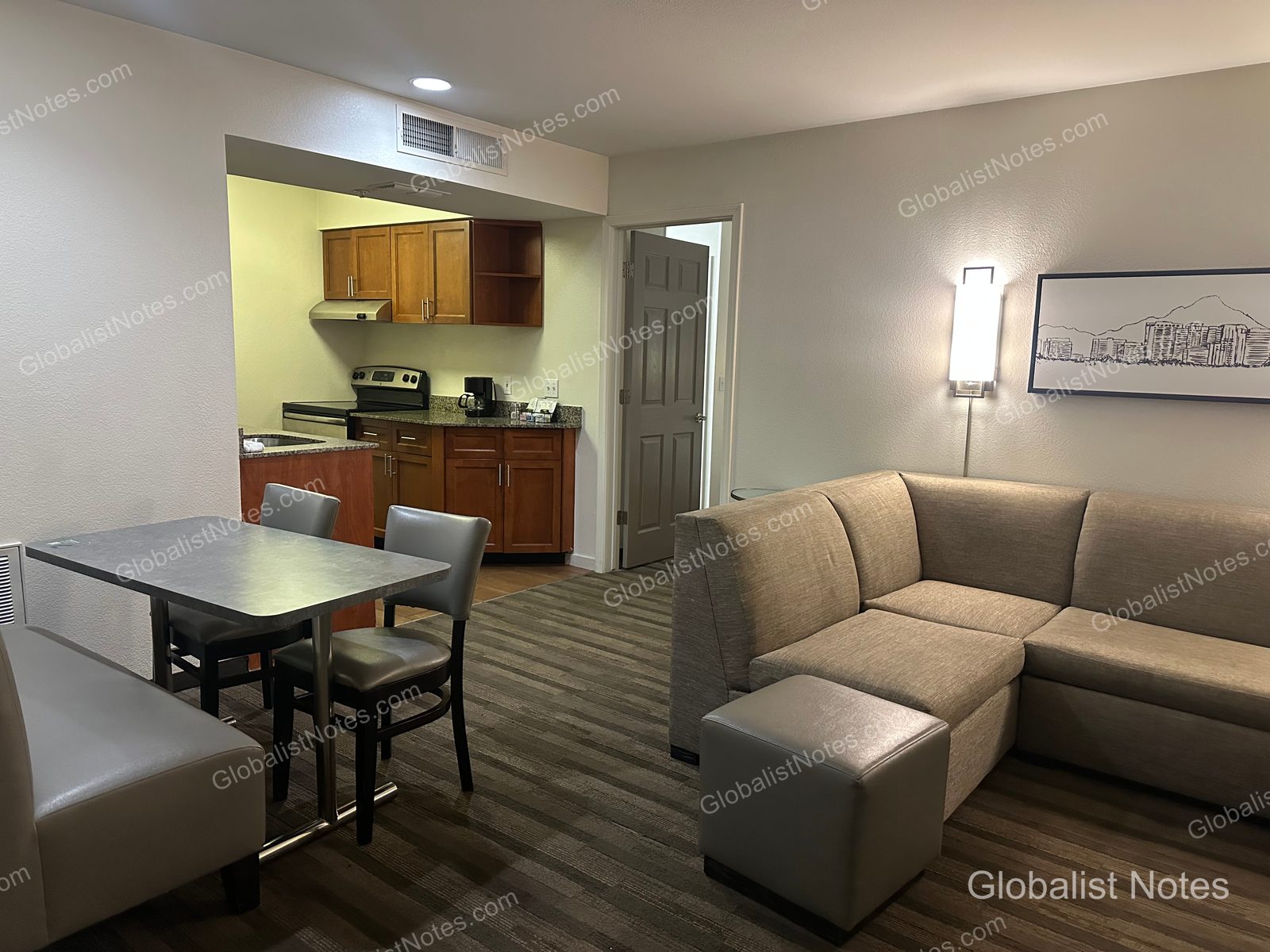 Hyatt House Scottsdale/Old Townの1ベッドルームスイートのリビングエリア。左手にダイニングテーブル、右手にグレーのコーナーソファとオットマンが配されています。奥にはキッチンが見え、壁にはスコッツデールの風景写真が飾られています。