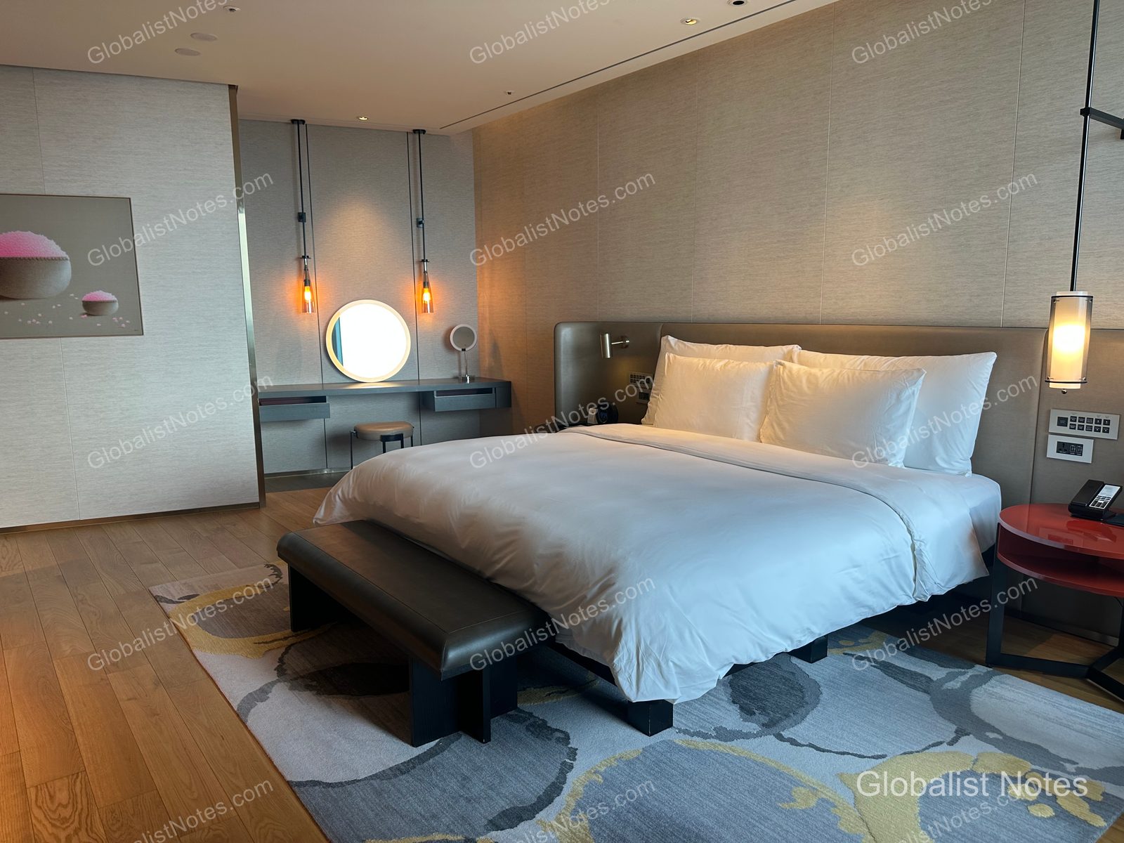Bedroom view of a One-Bedroom Suite at Grand Hyatt Jeju. A leather bench is placed at the foot of the king bed, and a vanity area with a circular mirror and lighting is set up beside the bed, over an abstract-patterned carpet.