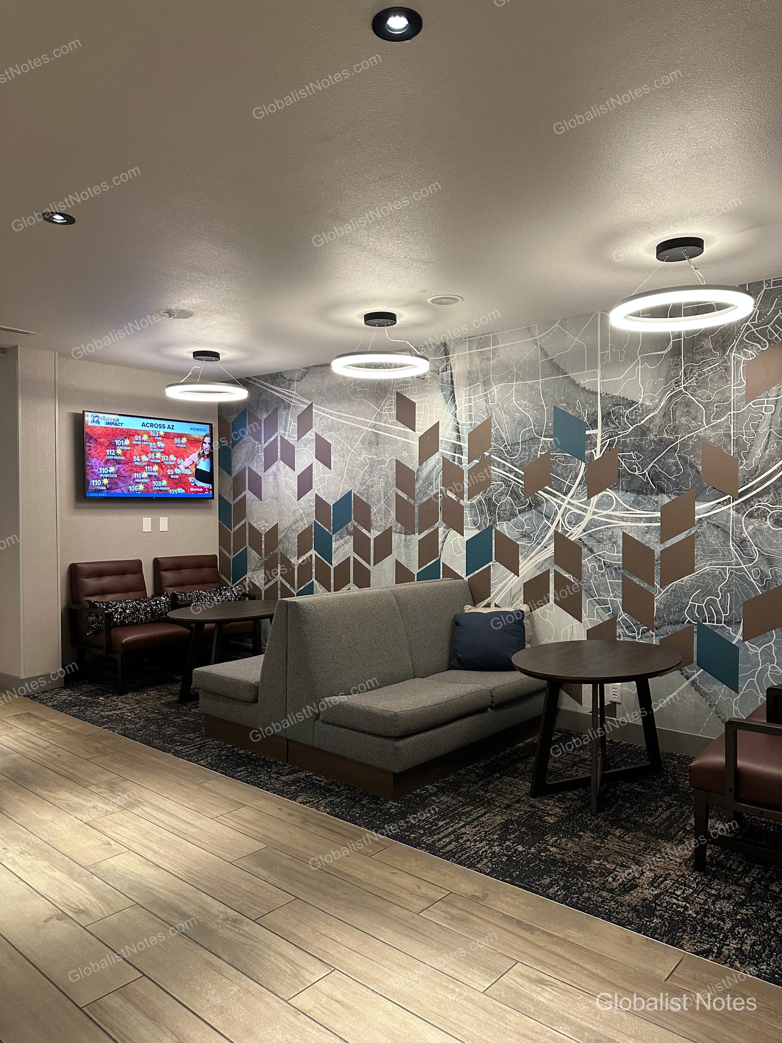 Hyatt Place Flagstaffのロビーラウンジ。壁面にはフラッグスタッフの地図をあしらったグラフィックアートが飾られ、手前にはグレーのソファ、ブラウンのレザーチェア、円形テーブルが配されています。天井にはモダンな円形照明が下がり、壁のモニターにはアリゾナ州の天気予報が映し出されています。