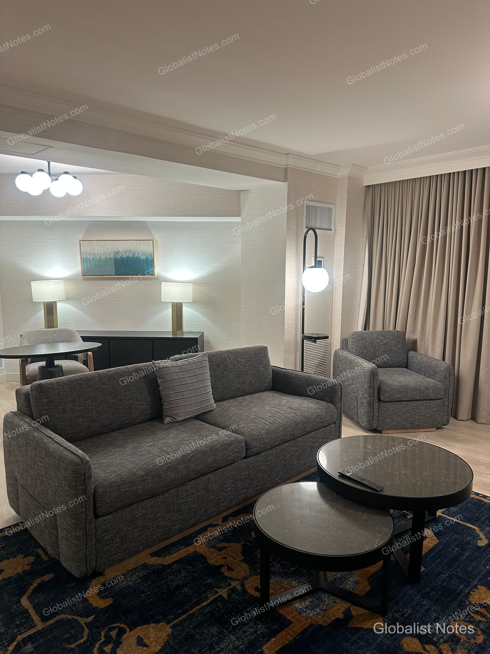 Hyatt Regency Irvine 一房套房客廳，配置深灰色沙發、單人扶手椅及兩張圓形咖啡桌，後方設有餐桌與玄關桌，地面鋪設深藍色花紋地毯。