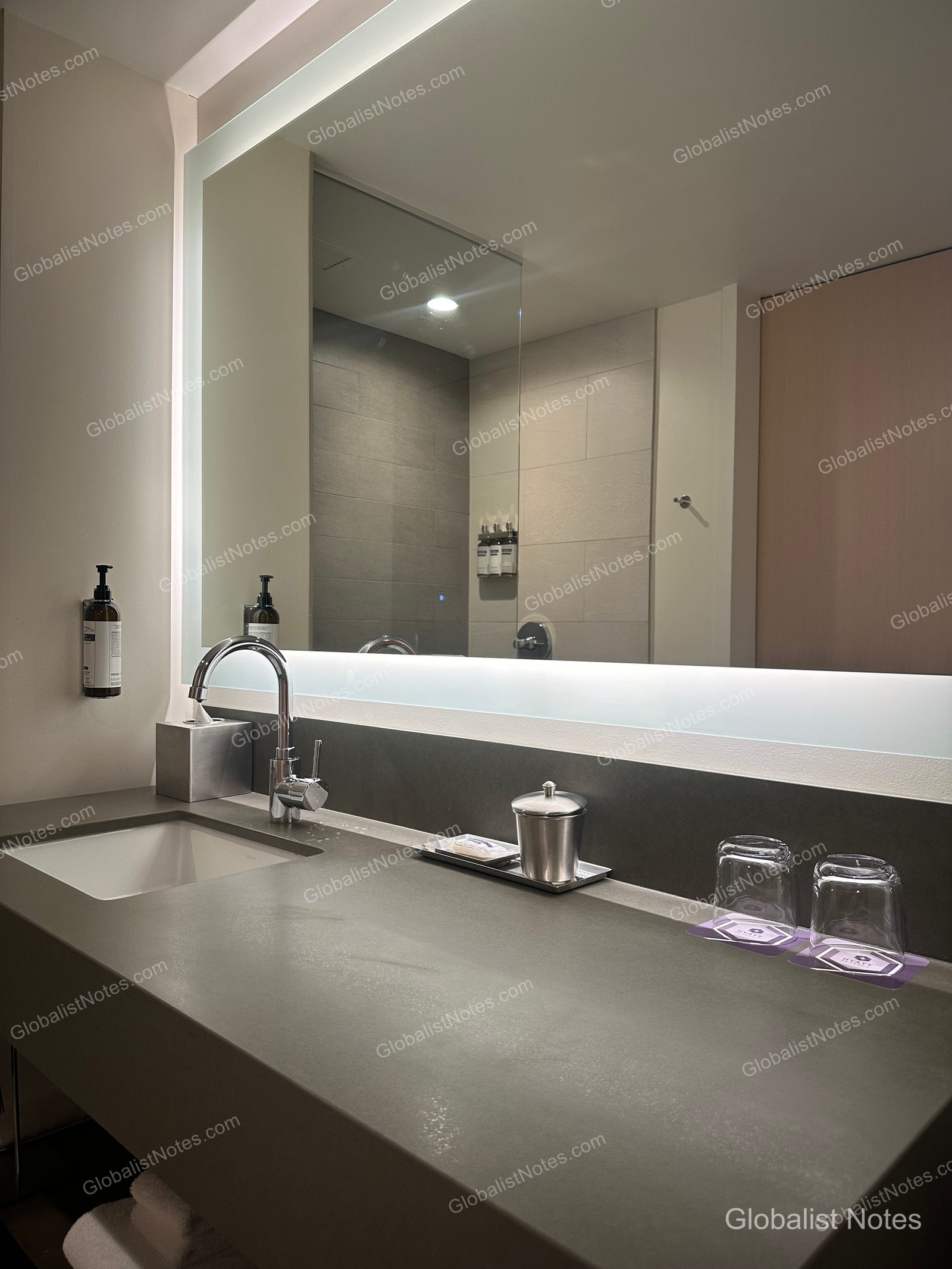 Vanity area in a Standard King room bathroom at Hyatt Regency Seattle. A long, gray-toned countertop features a large mirror with soft, indirect lighting, and the wall is neatly organized with amenities, faucets, and glassware.