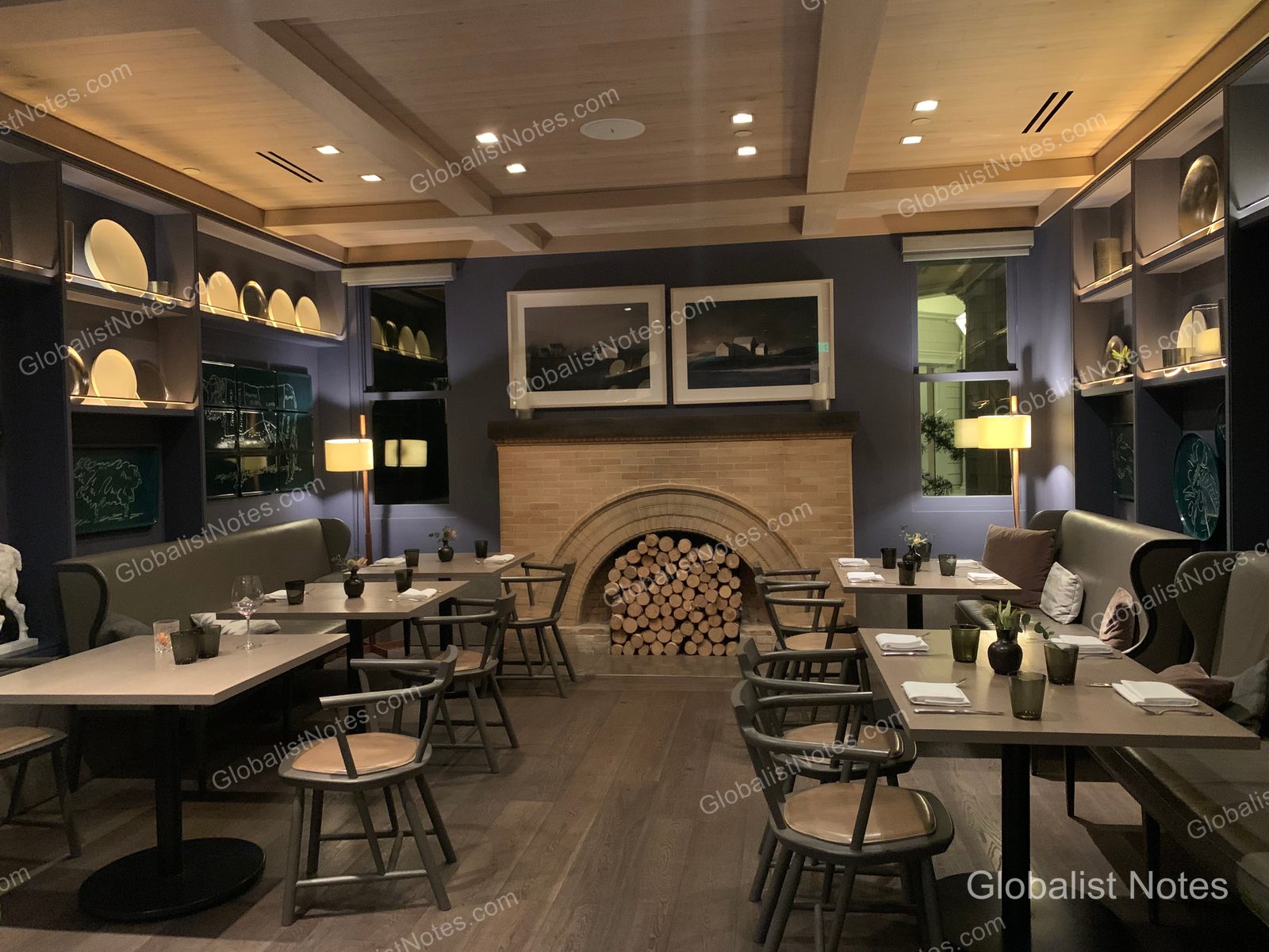 Indoor dining area at Alila Napa Valley in St. Helena, USA. An arched fireplace stacked with firewood serves as the centerpiece, flanked by neatly arranged tables and chairs. Decorative plates line the wall shelves, and the space is illuminated by soft, ambient lighting.