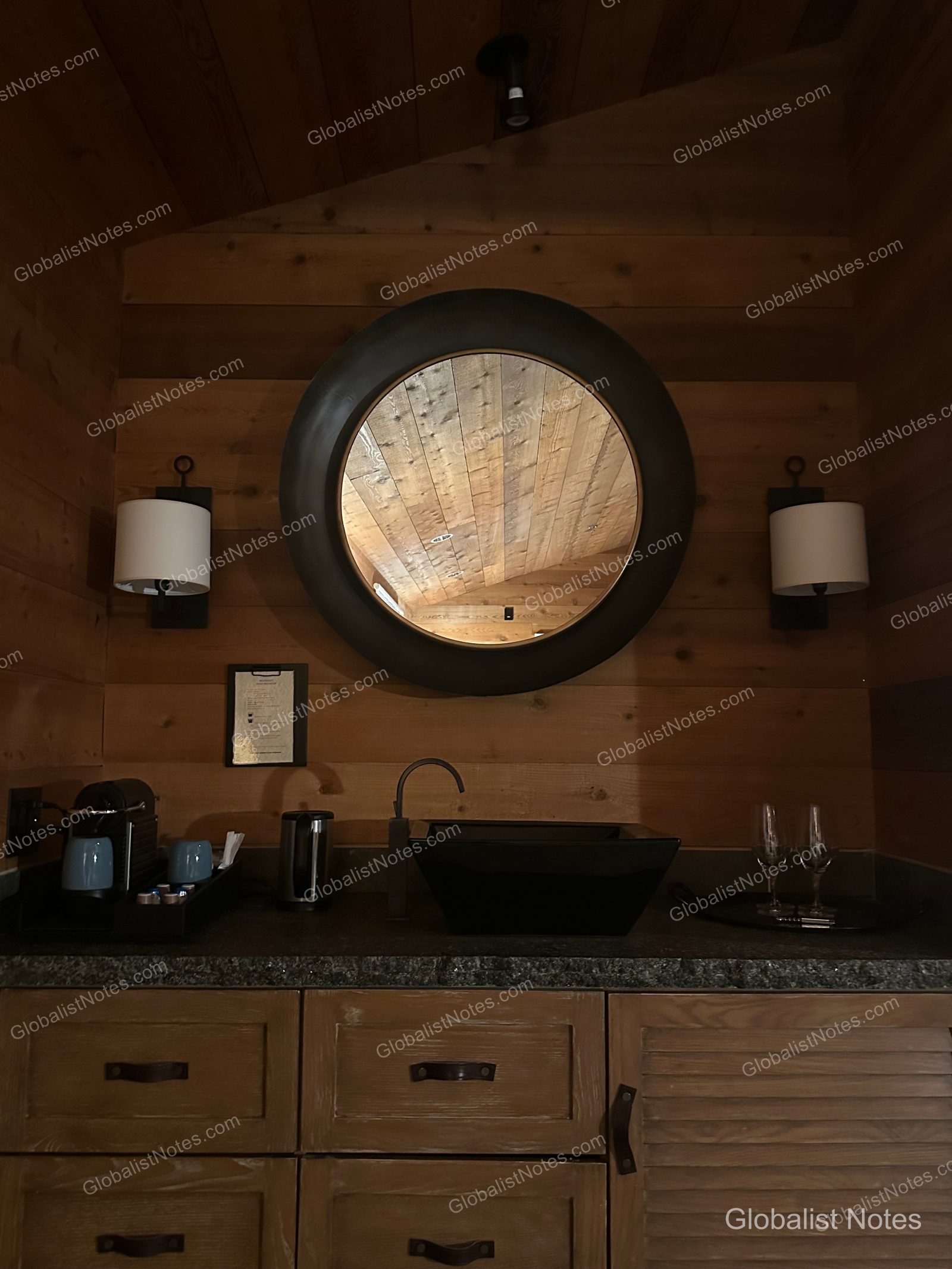 A vanity area in an Alila Ventana Big Sur Junior Suite featuring a round mirror and black square sink on a wooden wall, with wall sconces, a coffee machine, and wine glasses.