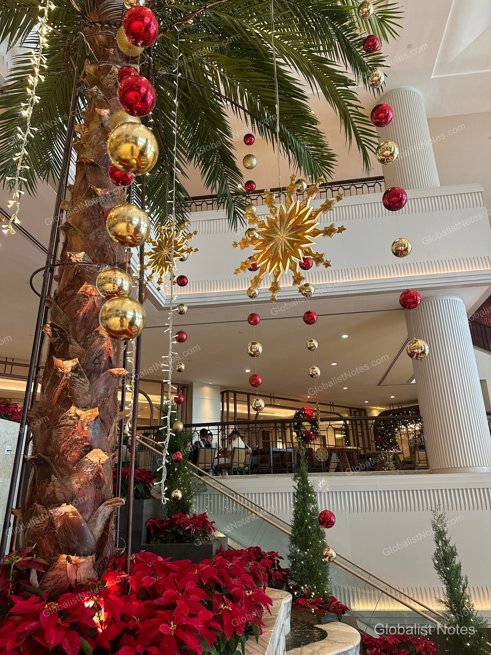 Christmas decorations in the Grand Hyatt Jakarta lobby, featuring gold and red spherical ornaments hanging along palm tree trunks, surrounded by poinsettias and small Christmas trees, with the hotel's interior staircase and railings visible in the background.