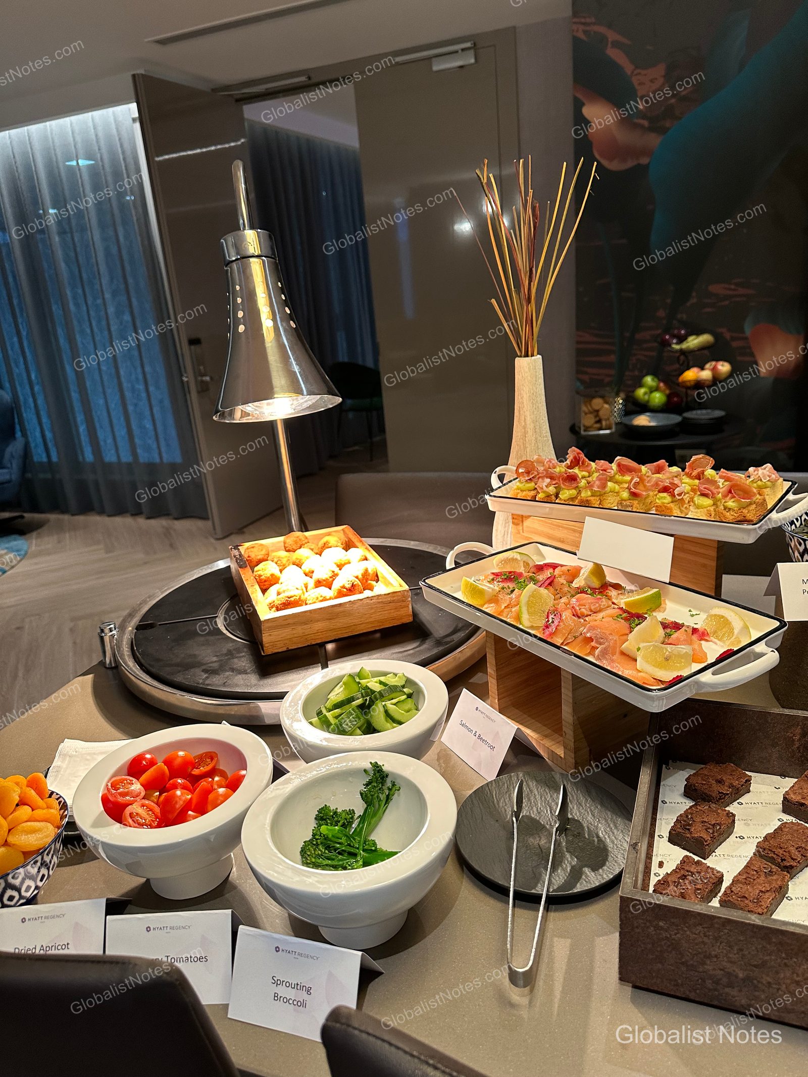 A buffet spread in the Hyatt Regency Malta lounge featuring salmon, broccoli, cherry tomatoes, dried apricots, and brownies on white plates and in wooden crates under warm lighting.