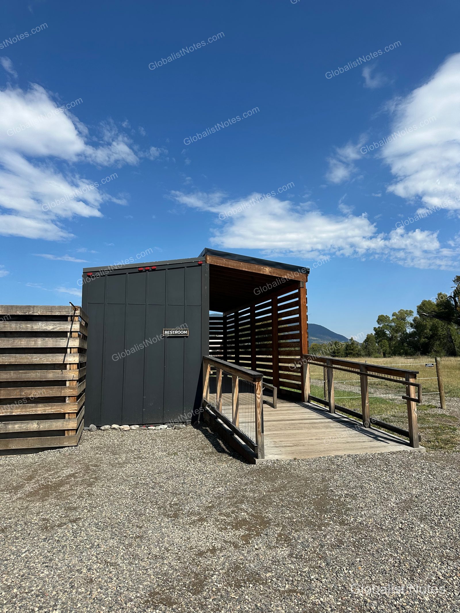 Under Canvas North Yellowstoneの共用トイレ棟。黒い外壁の建物の横に木製スロープが設置され、周囲には砂利道と青空が広がっています。