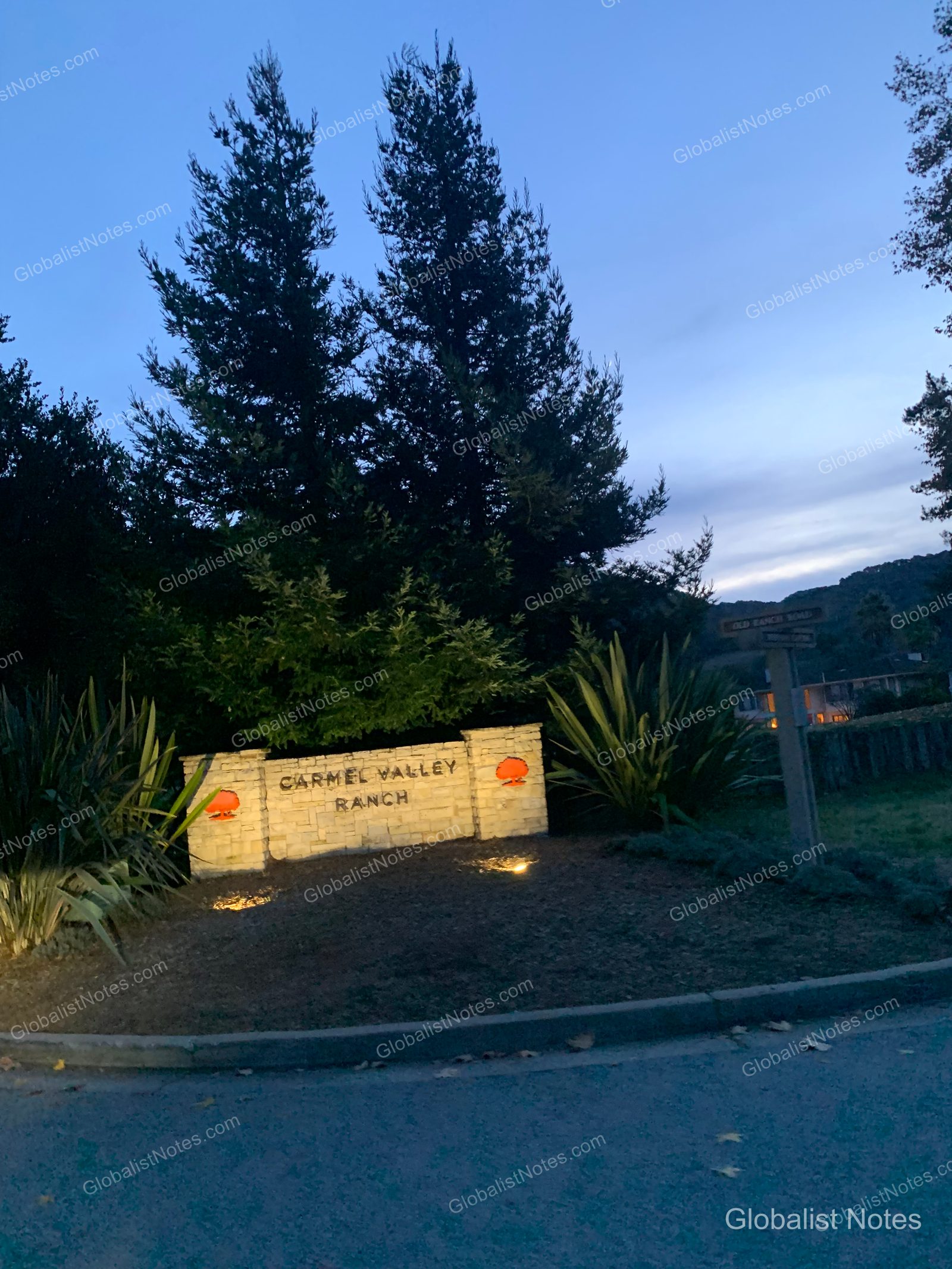 夕暮れ時、ライトアップされたCarmel Valley Ranchの石造りのエントランスサイン。周囲には背の高い木々と庭園が広がっています。