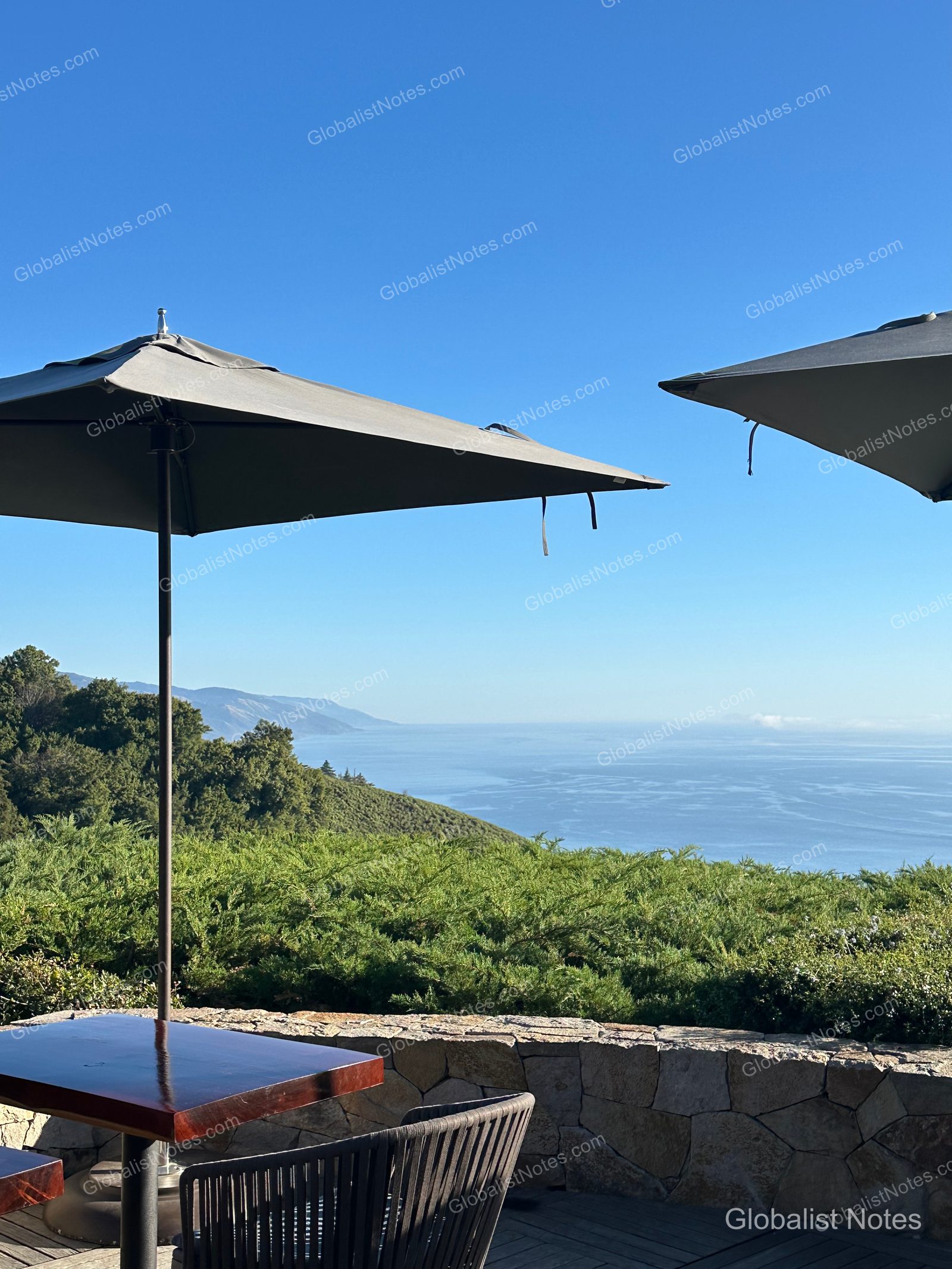 A view from the outdoor terrace at Alila Ventana Big Sur, featuring the Pacific coastline and cliffs under dark gray umbrellas, with stone walls and wooden chairs.