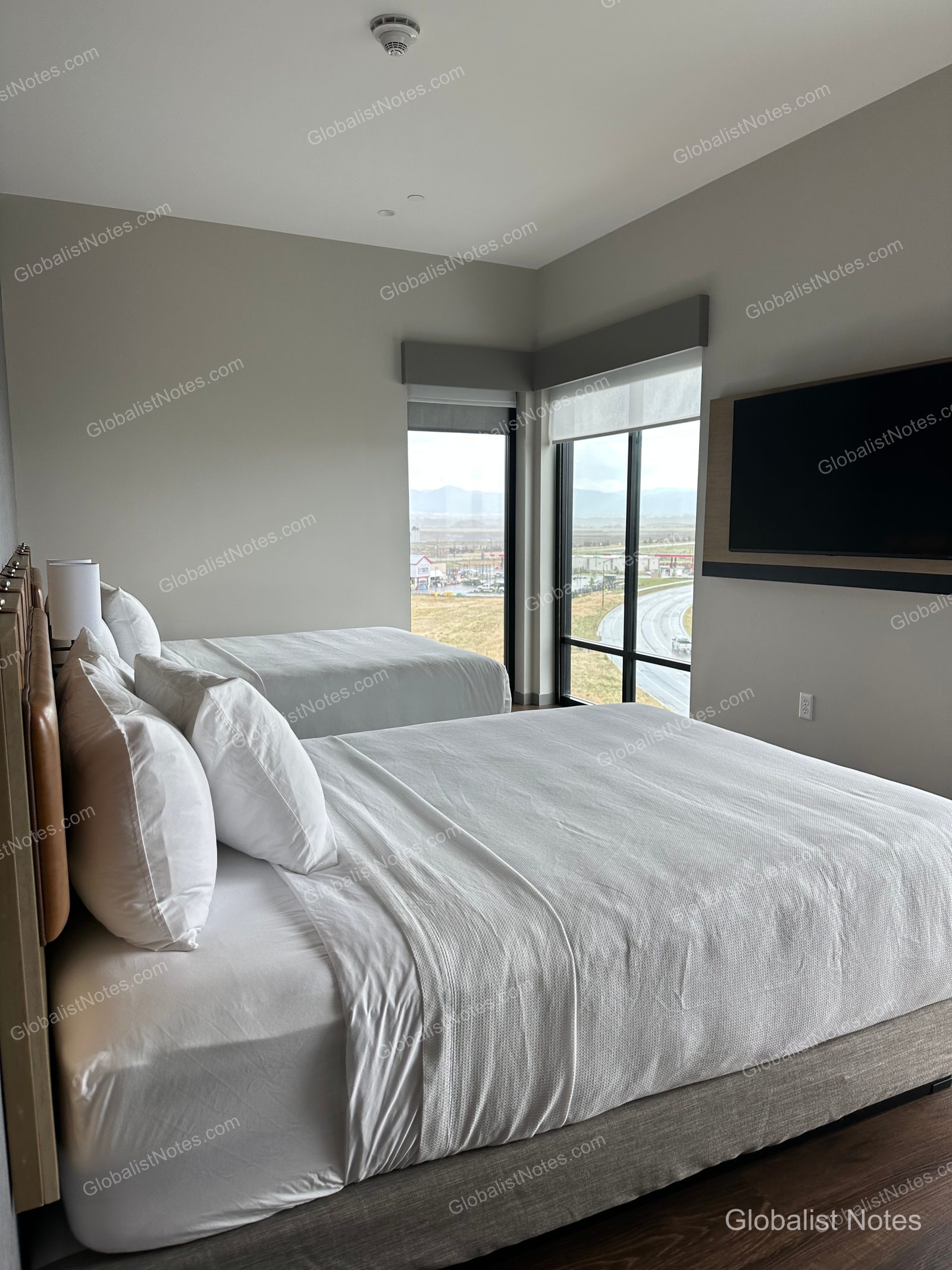 Hyatt Place Bozeman Yellowstone Airportのジュニアスイートの室内。ベッド2台が並び、窓からはBelgradeの道路と平原が見渡せます。壁面にはテレビが設置されています。