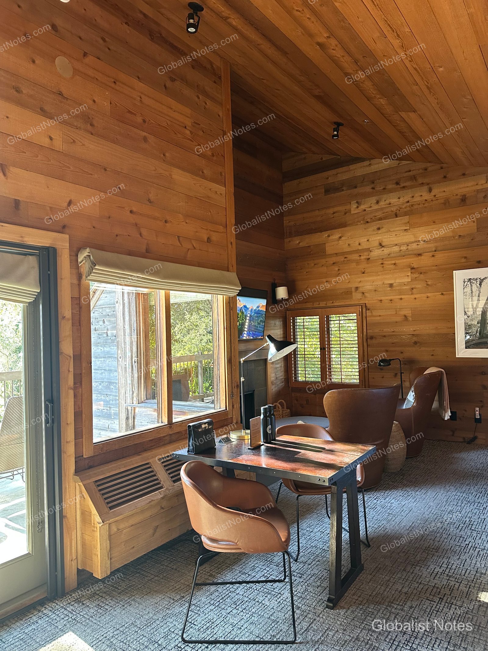 Interior of an Alila Ventana Big Sur Junior Suite, featuring a cozy living area with a table, leather chairs, wood-paneled walls, high ceilings, and a view of the terrace through the window.
