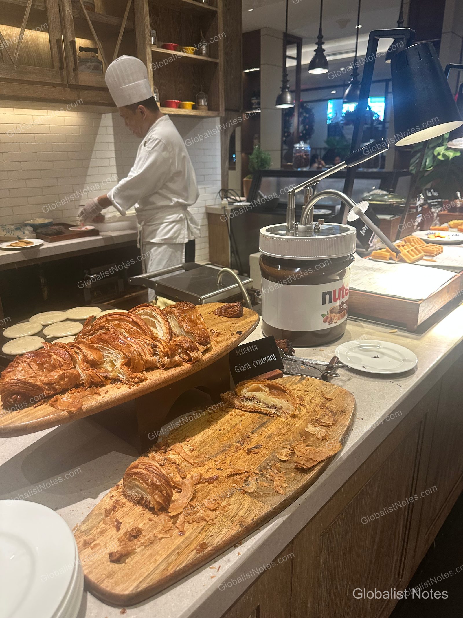 Bakery section of the Grand Hyatt Jakarta breakfast buffet, featuring a large Nutella croissant on a wooden board next to a large-capacity Nutella dispenser. In the background, a chef prepares food at a station, with various breakfast items like waffles and pancakes displayed nearby.