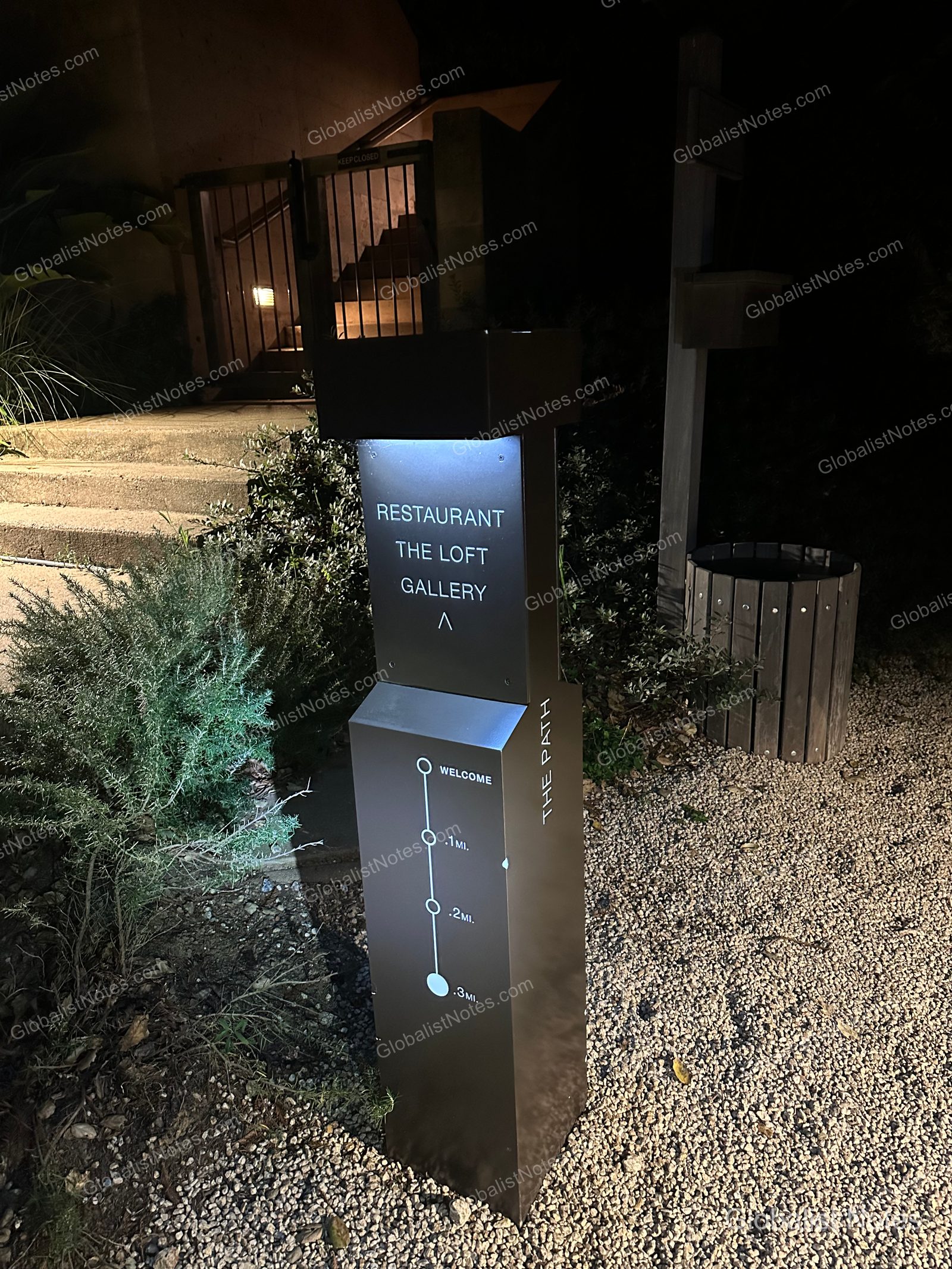 A metal signpost at Alila Ventana Big Sur at night, showing directions and distances (0.1 to 0.3 miles) to the restaurant, The Loft, and the gallery.