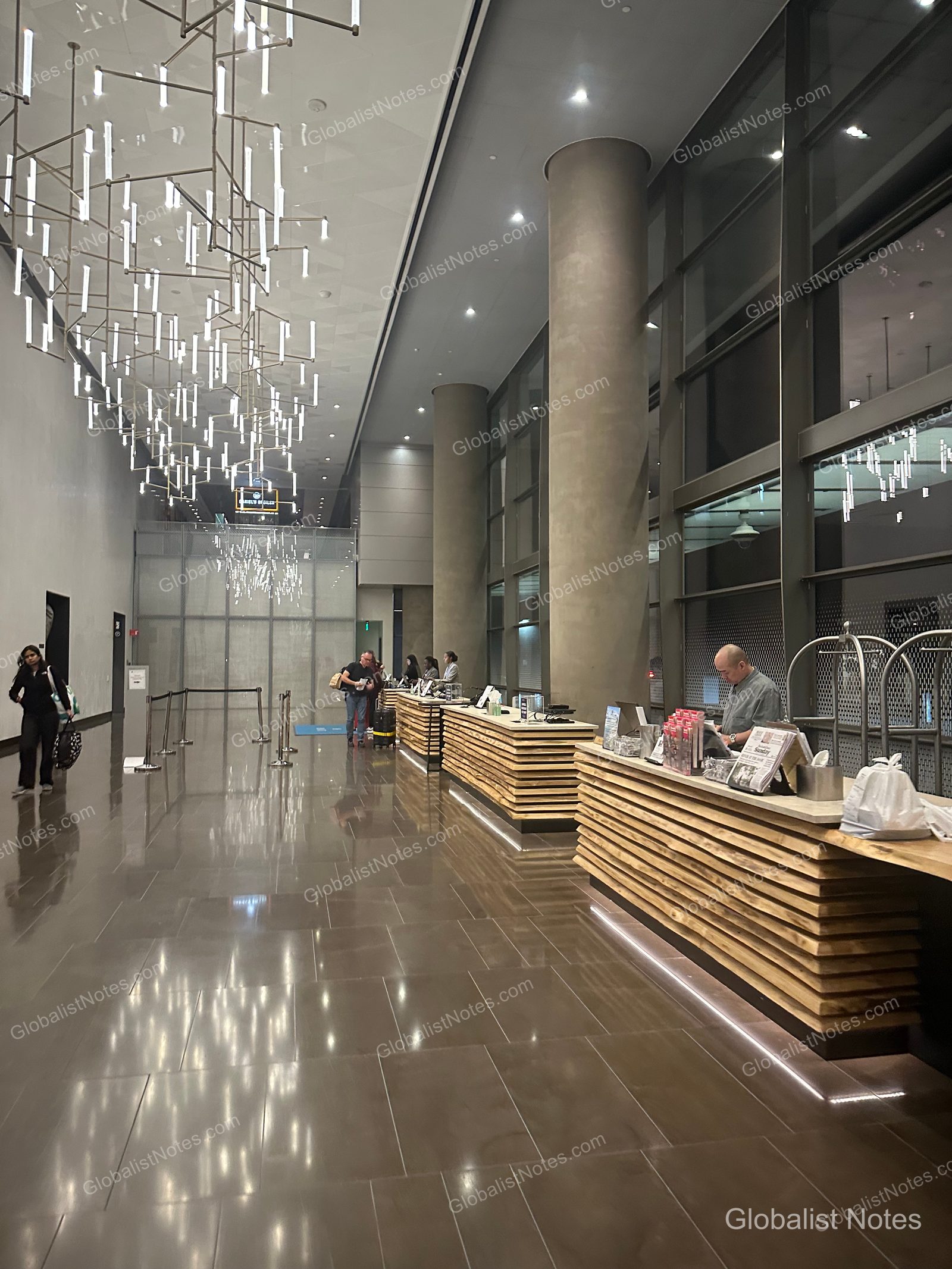 Modern lobby view at Hyatt Regency Seattle. The high ceiling features numerous hanging rod-style light fixtures, leading to a long wood-slab check-in counter and marble flooring. Guests are seen checking in at the front desk.