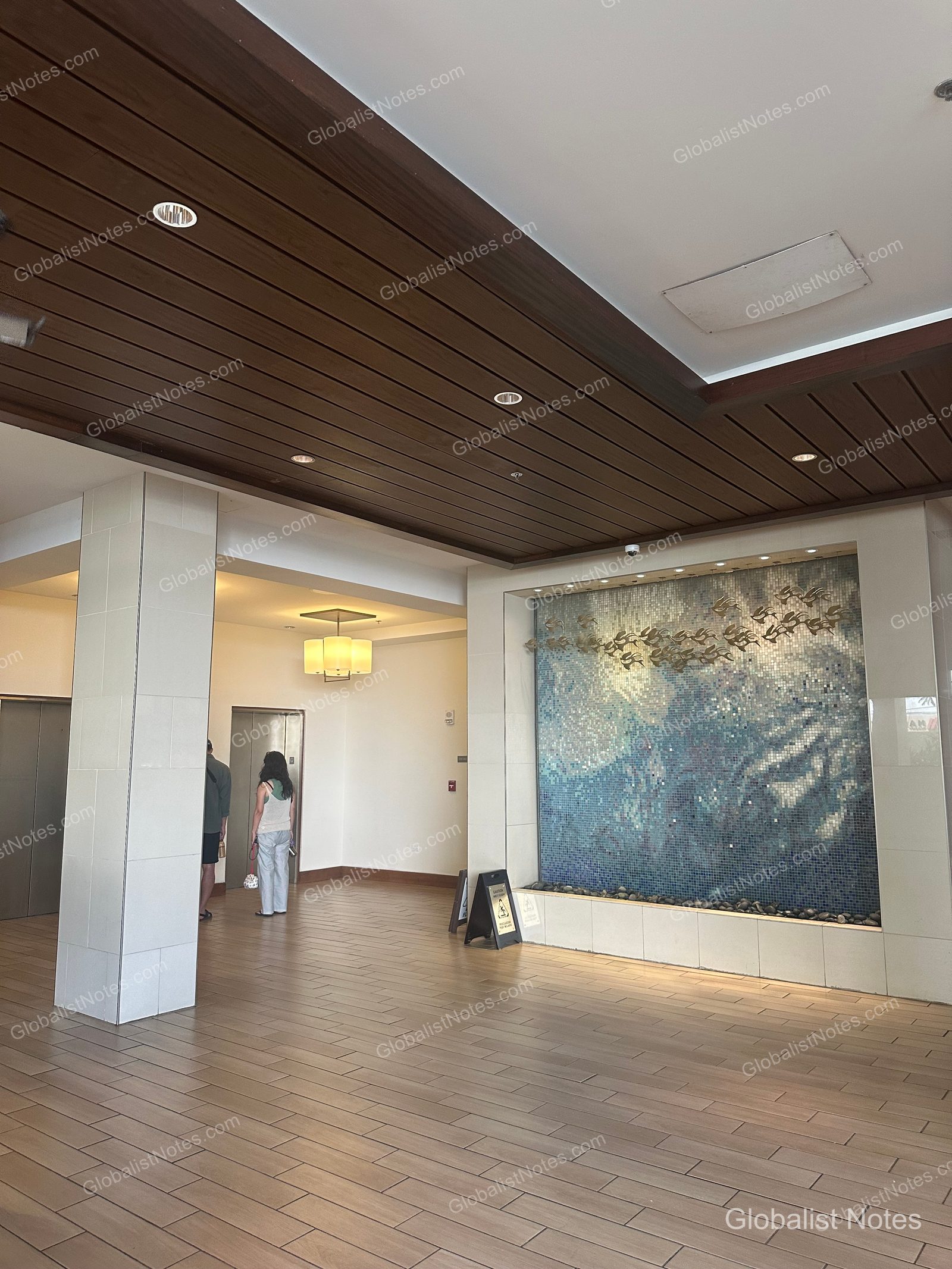 Lobby view of the Hyatt Place Waikiki Beach in Honolulu, Hawaii. The space features a wood-textured ceiling and light tile floors, with a water feature decorated with blue mosaic tiles and metal accents on one wall, and guests waiting for the elevator nearby.