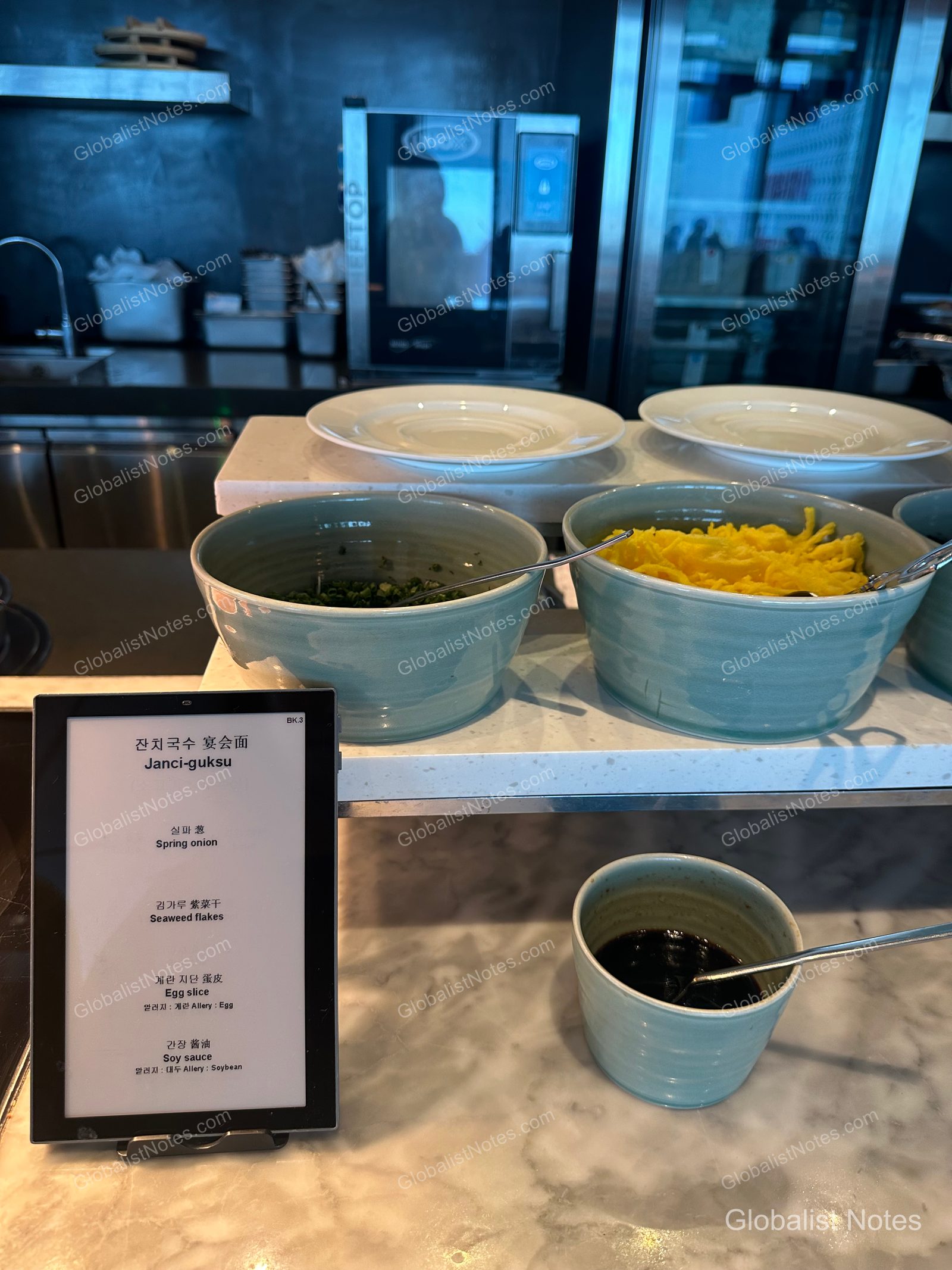 Janchi-guksu (noodle soup) station at the Grand Hyatt Jeju breakfast buffet. Ceramic bowls containing toppings like chopped green onions, dried seaweed, and egg garnish are neatly arranged on a marble table alongside soy sauce and a menu sign.