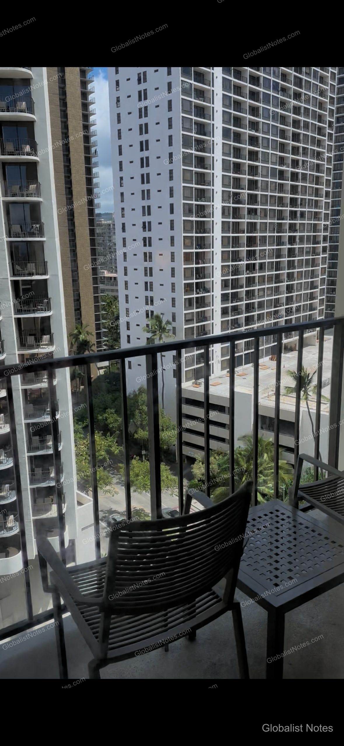 View from the balcony of a guest room at Hyatt Place Waikiki Beach in Honolulu, Hawaii. The balcony features a black metal railing, a small table, and chairs, looking out over nearby high-rise buildings, palm trees, and the street below.