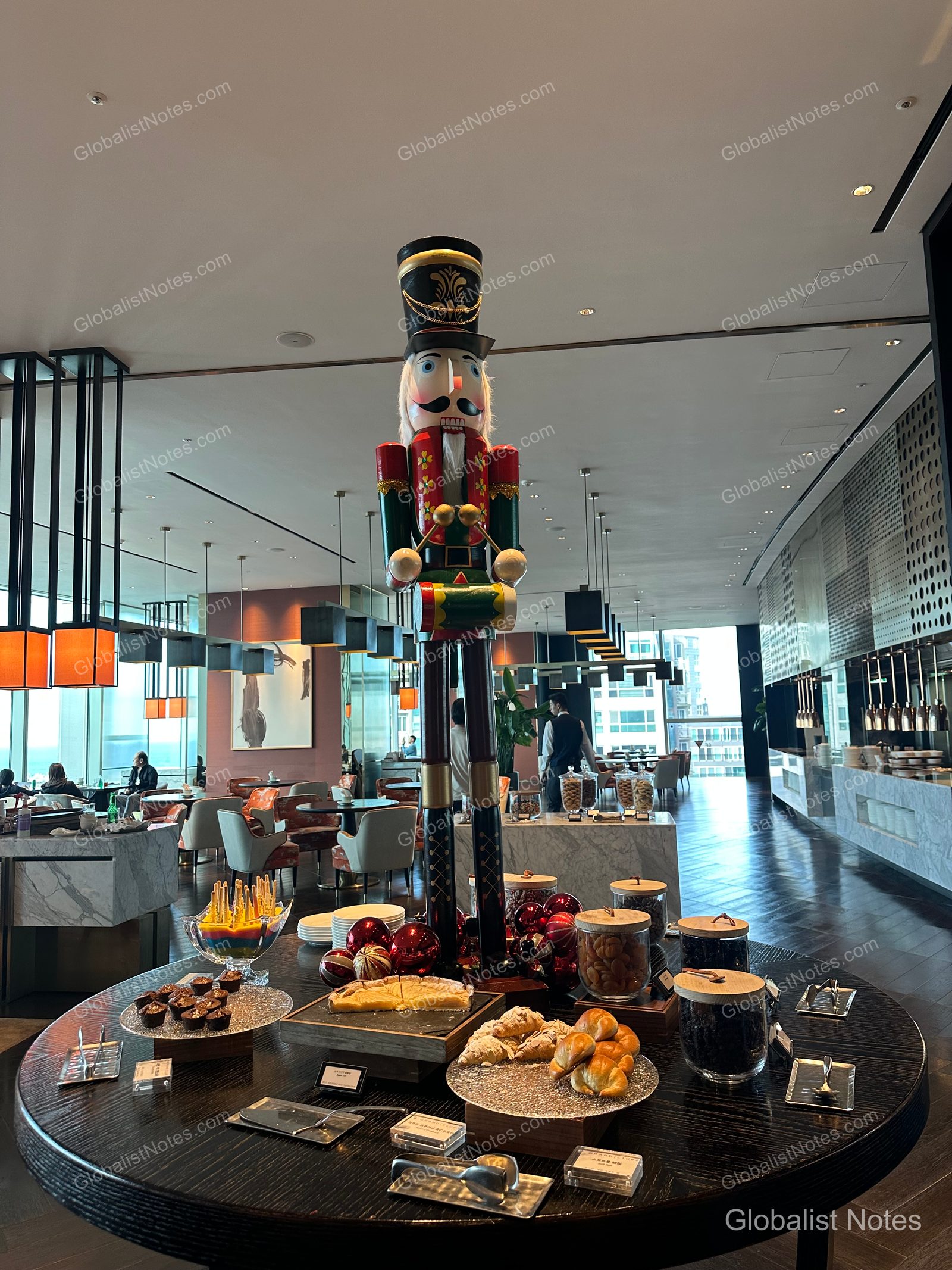 A large nutcracker decoration stands on a circular table in the center of the Grand Hyatt Jeju lounge, surrounded by an assortment of desserts and pastries. Floor-to-ceiling windows offer a view of Jeju City, with lounge seating arranged throughout the space.