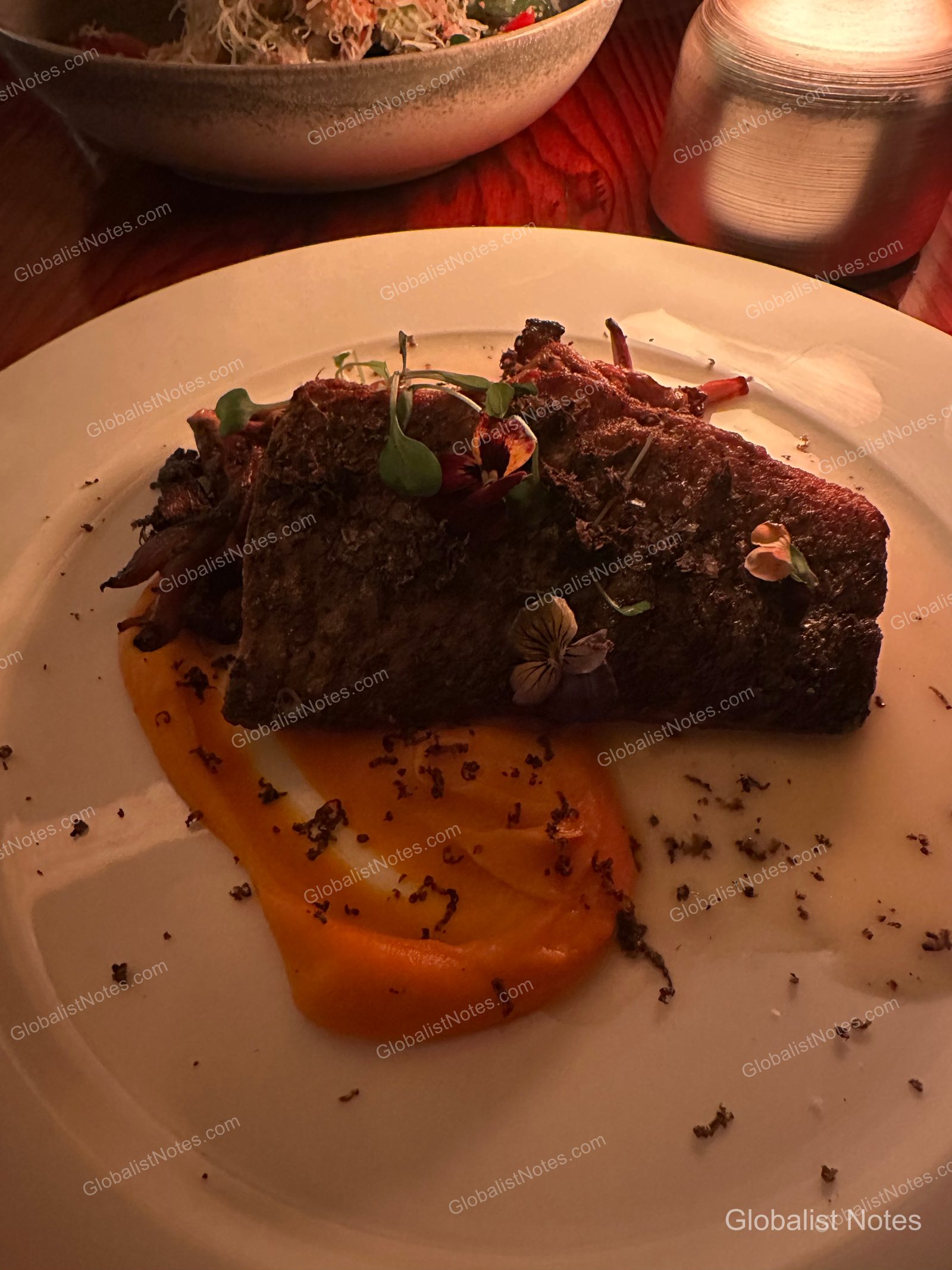 Dinner entree at Alila Ventana Big Sur featuring roasted meat served over an orange puree, garnished with edible flowers and herbs on a table in low lighting.
