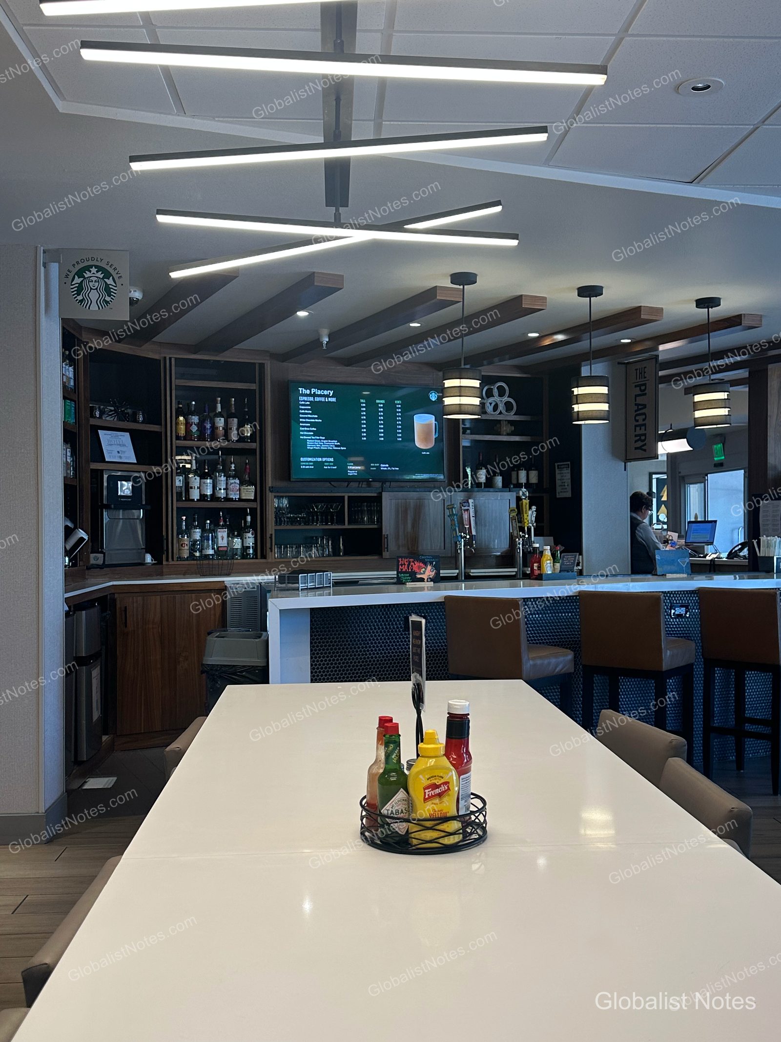 Hyatt Place Flagstaffの「The Placery」バー。手前の白いテーブルにはソース類が並び、奥にはバーカウンターとスターバックスのメニューモニター、モダンな照明が配されています。
