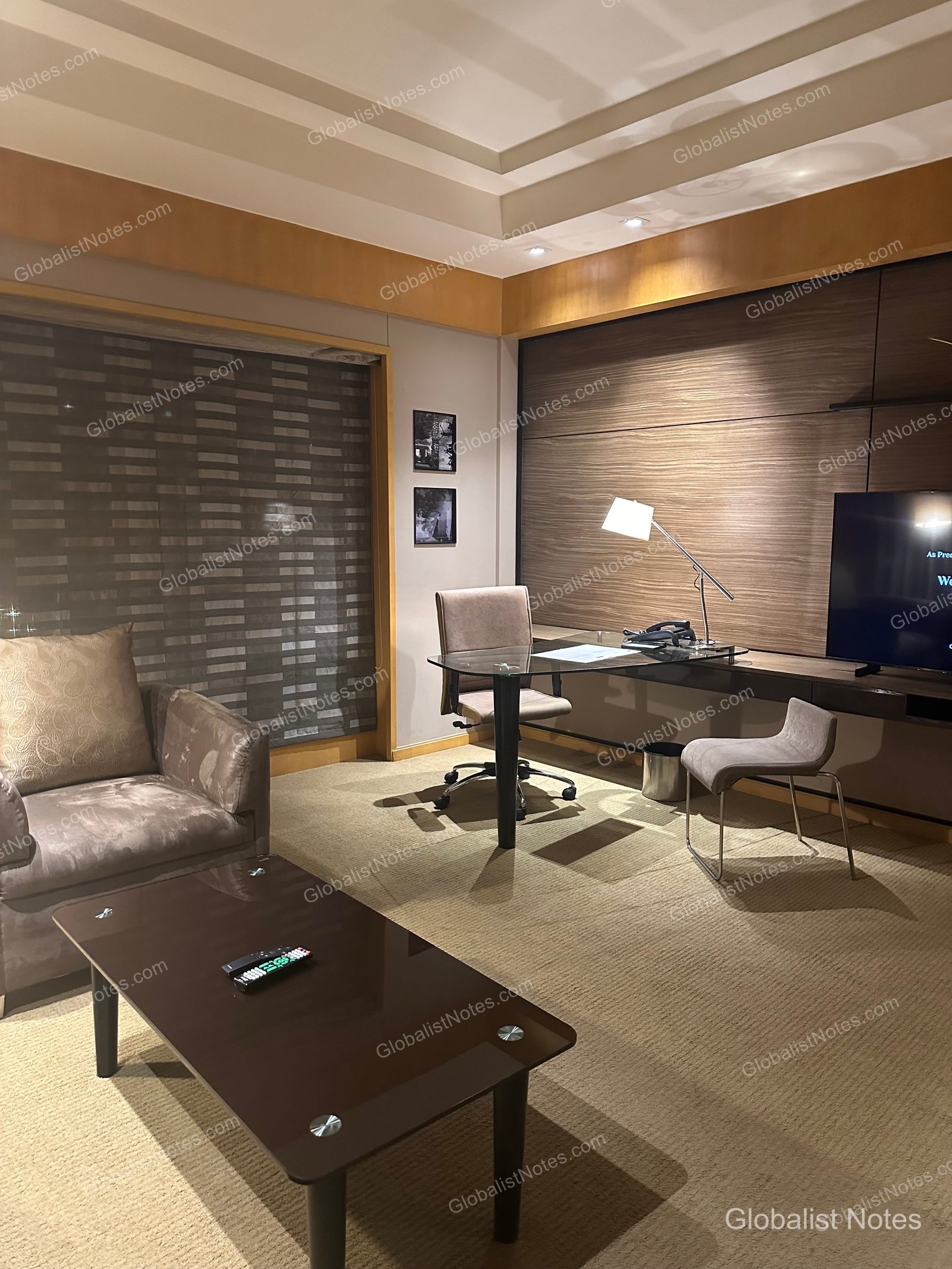 Interior view of the Veranda Suite King at Grand Hyatt Mumbai Hotel and Residences, showing a brown-toned wall, carpeted flooring, a glass-topped desk, chair, sofa, and low table.