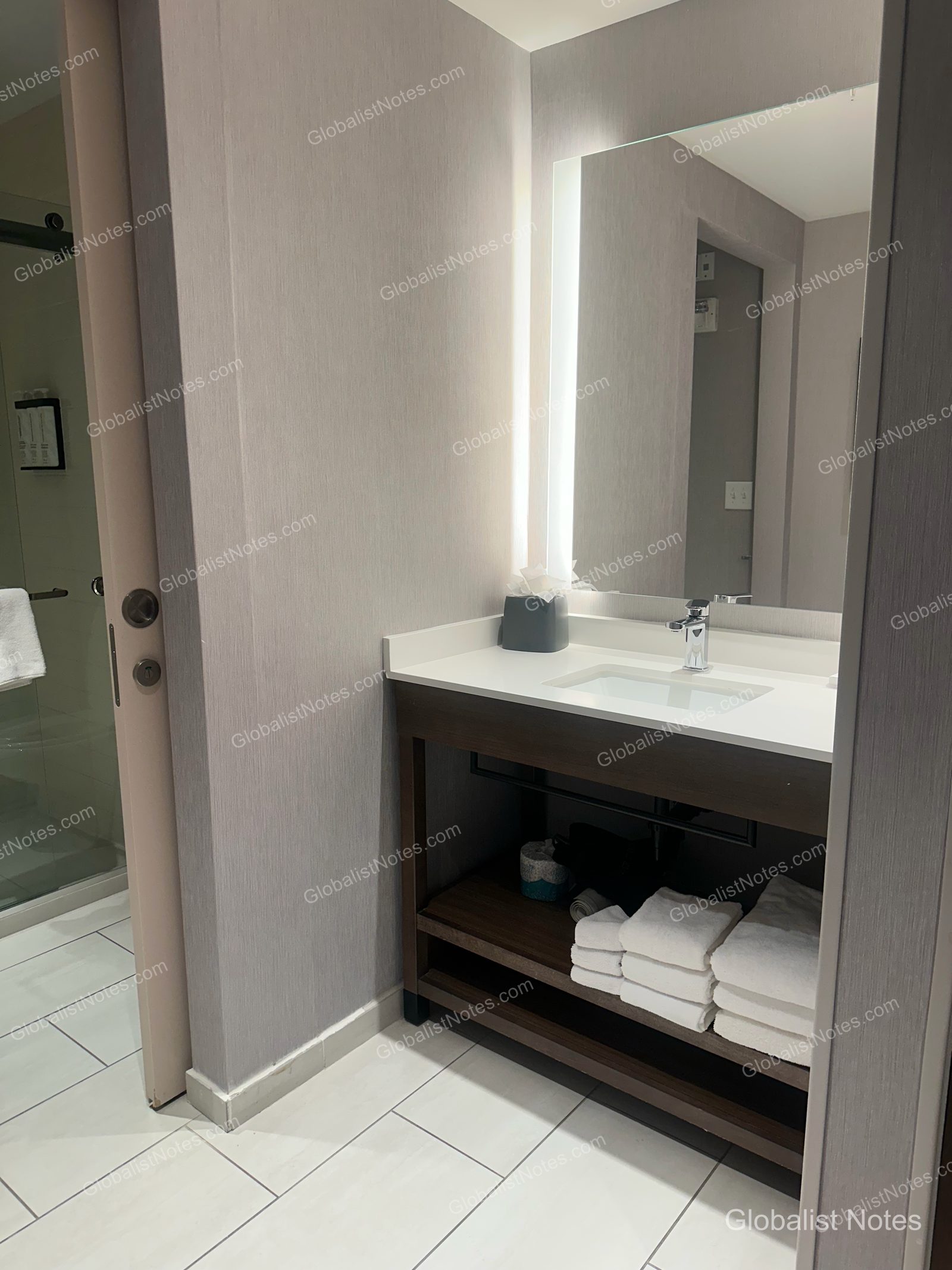 Hyatt Place Flagstaffのデラックスルームのバスルーム。明るい鏡と白い洗面台、下段の棚にはタオルが整然と並べられており、左側にはガラス扉のシャワーブースがあります。