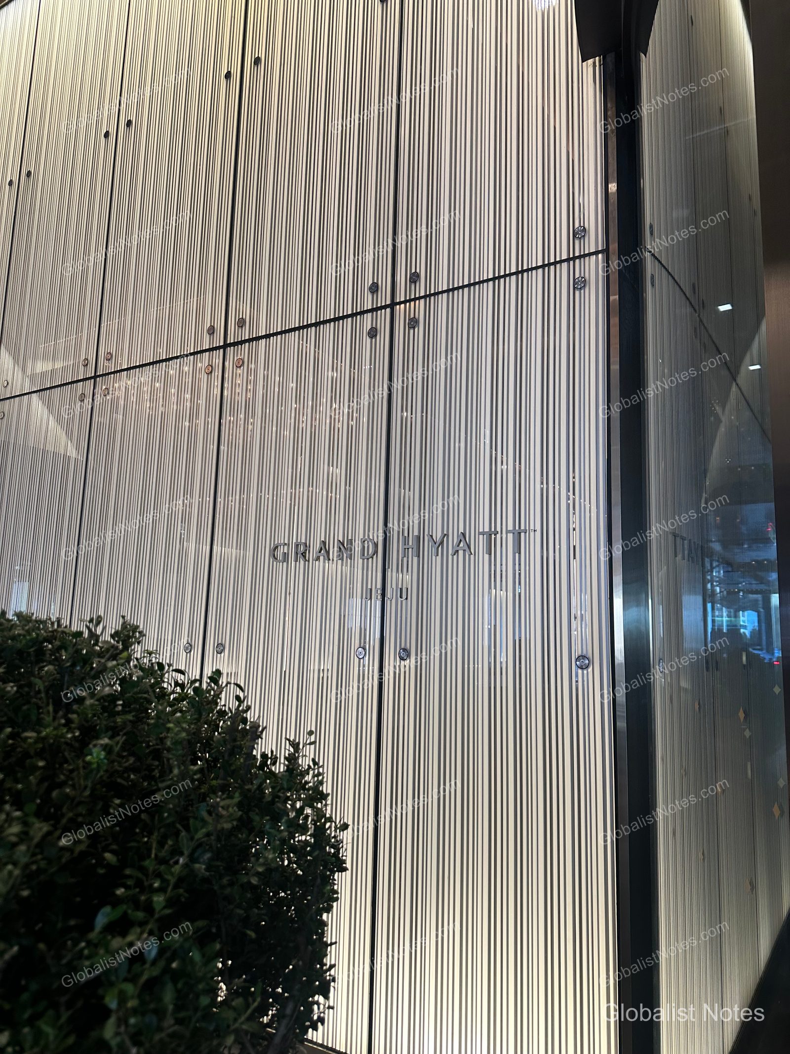 Exterior facade of Grand Hyatt Jeju, featuring densely arranged vertical metal panels illuminated by lights, with the Grand Hyatt logo centered. A portion of dark green shrubbery is visible in the bottom left corner.