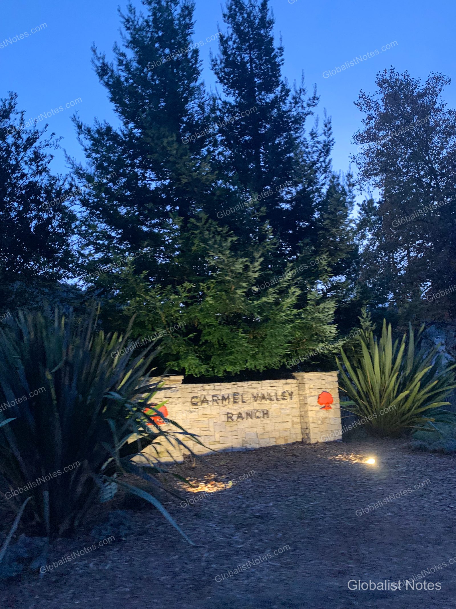夕暮れ時、ライトアップされたCarmel Valley Ranchの石造りのエントランスサイン。周囲には木々や植栽が広がっています。