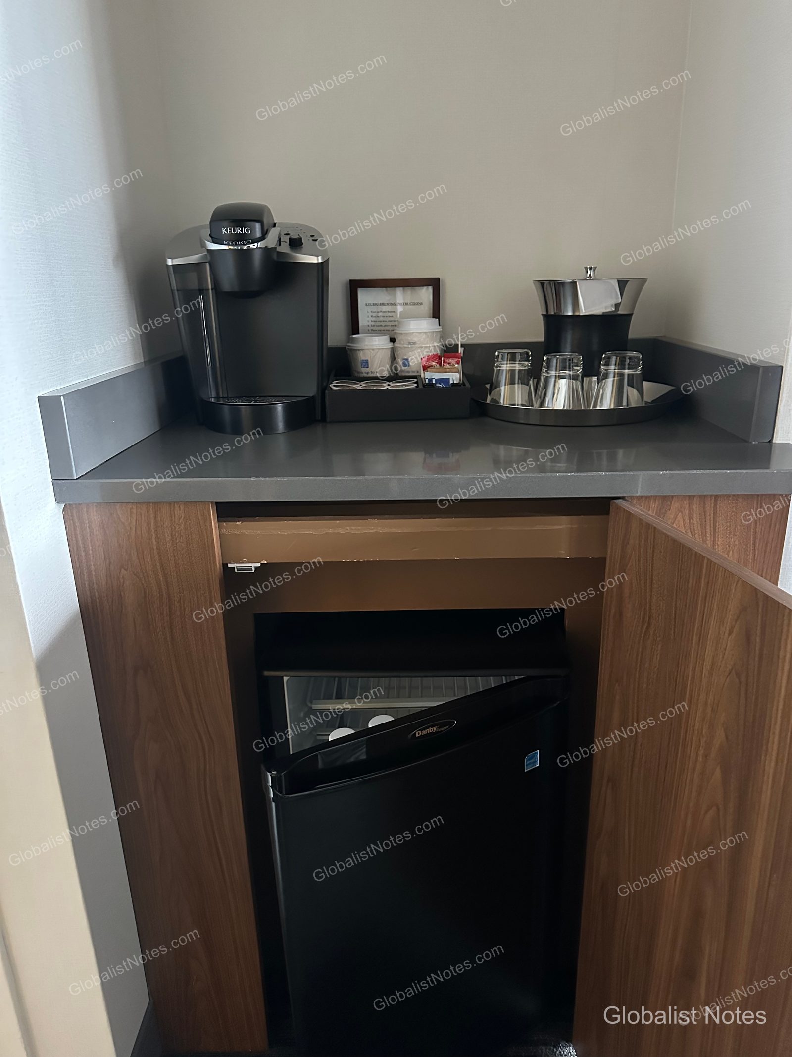 Minibar area in a Deluxe Suite at Hyatt Regency Vancouver. A black Keurig coffee machine, coffee pods, glassware, and an ice bucket are arranged on the grey countertop, with an empty Danby mini-fridge housed in the wooden cabinet below.