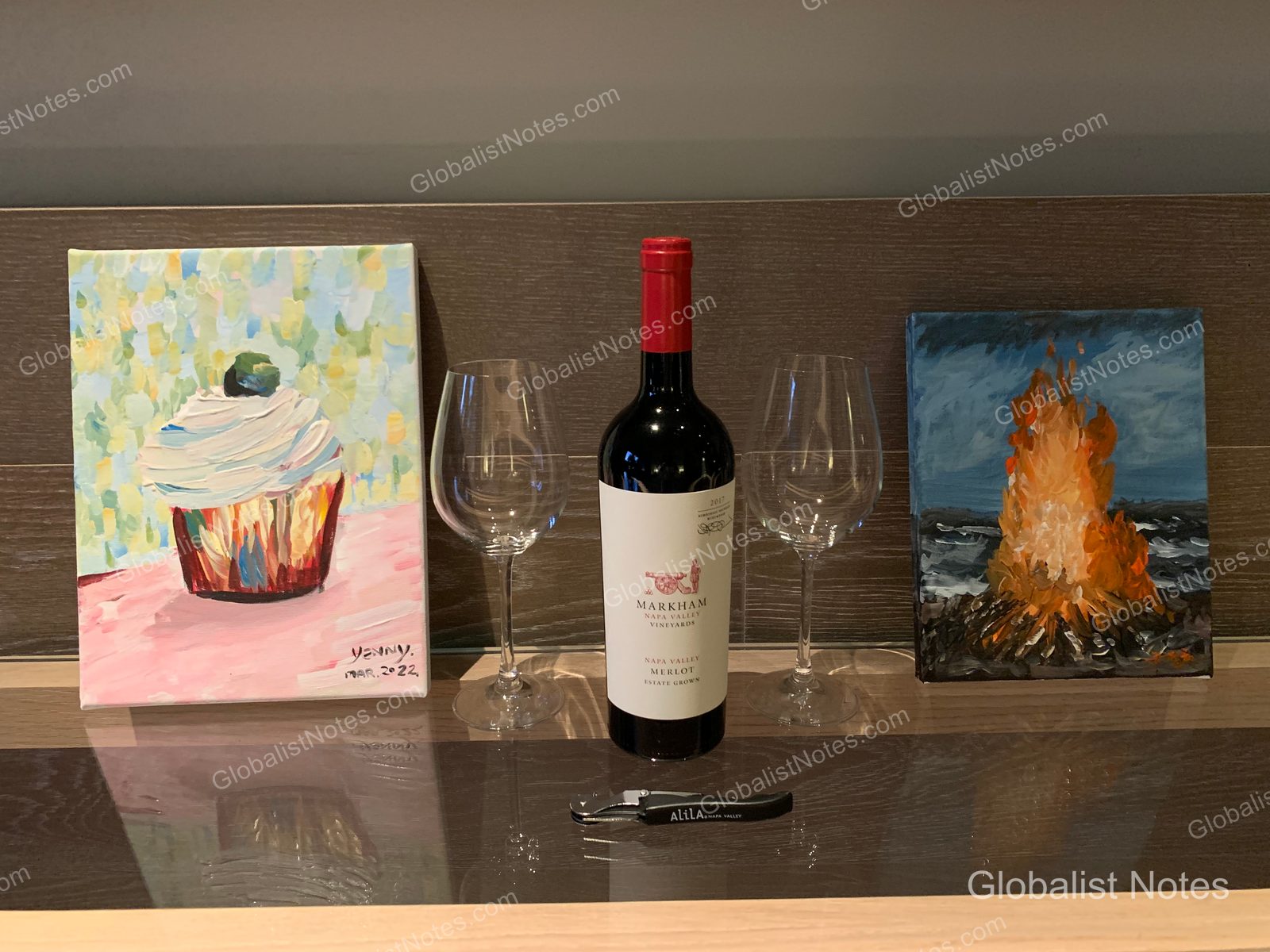 A bottle of Markham Merlot, two wine glasses, and a corkscrew set on the guest room table at Alila Napa Valley, flanked by two small canvas paintings depicting cupcakes and a campfire.