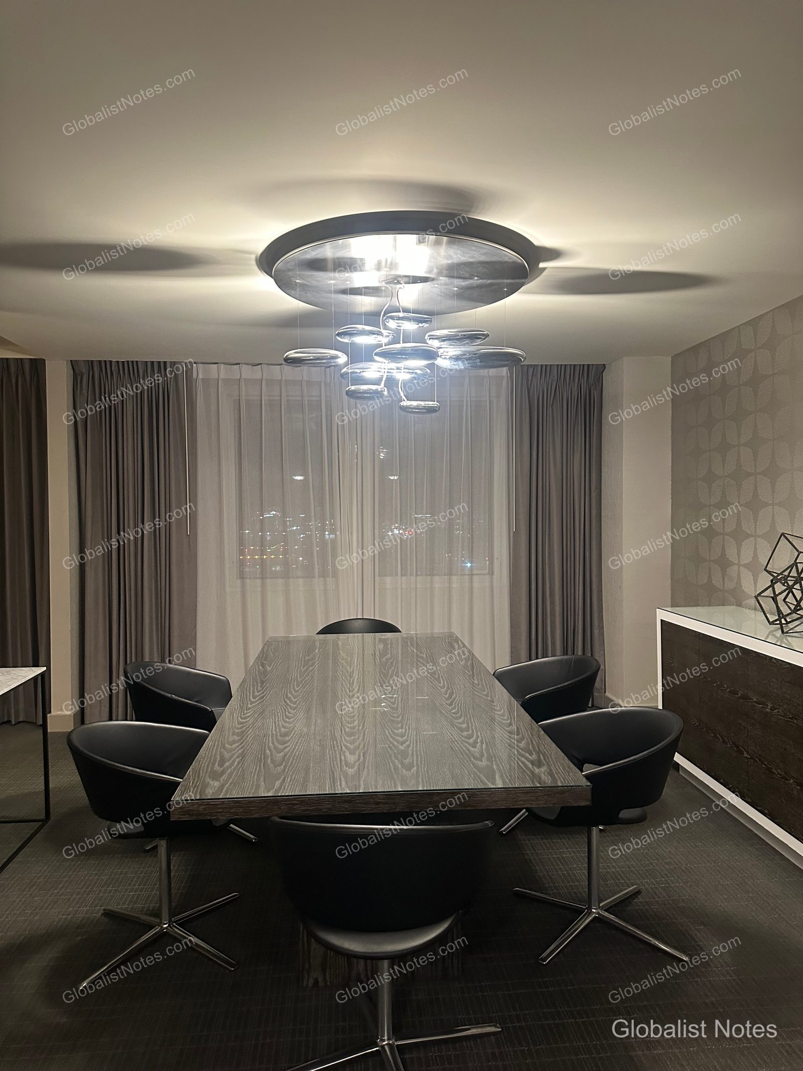 Dining area in a One Bedroom King Suite at Hyatt Regency Los Angeles International Airport. A long wooden table is surrounded by six black chairs, with a modern chandelier featuring multiple small lights overhead. Curtained windows and patterned wallpaper are visible in the background.