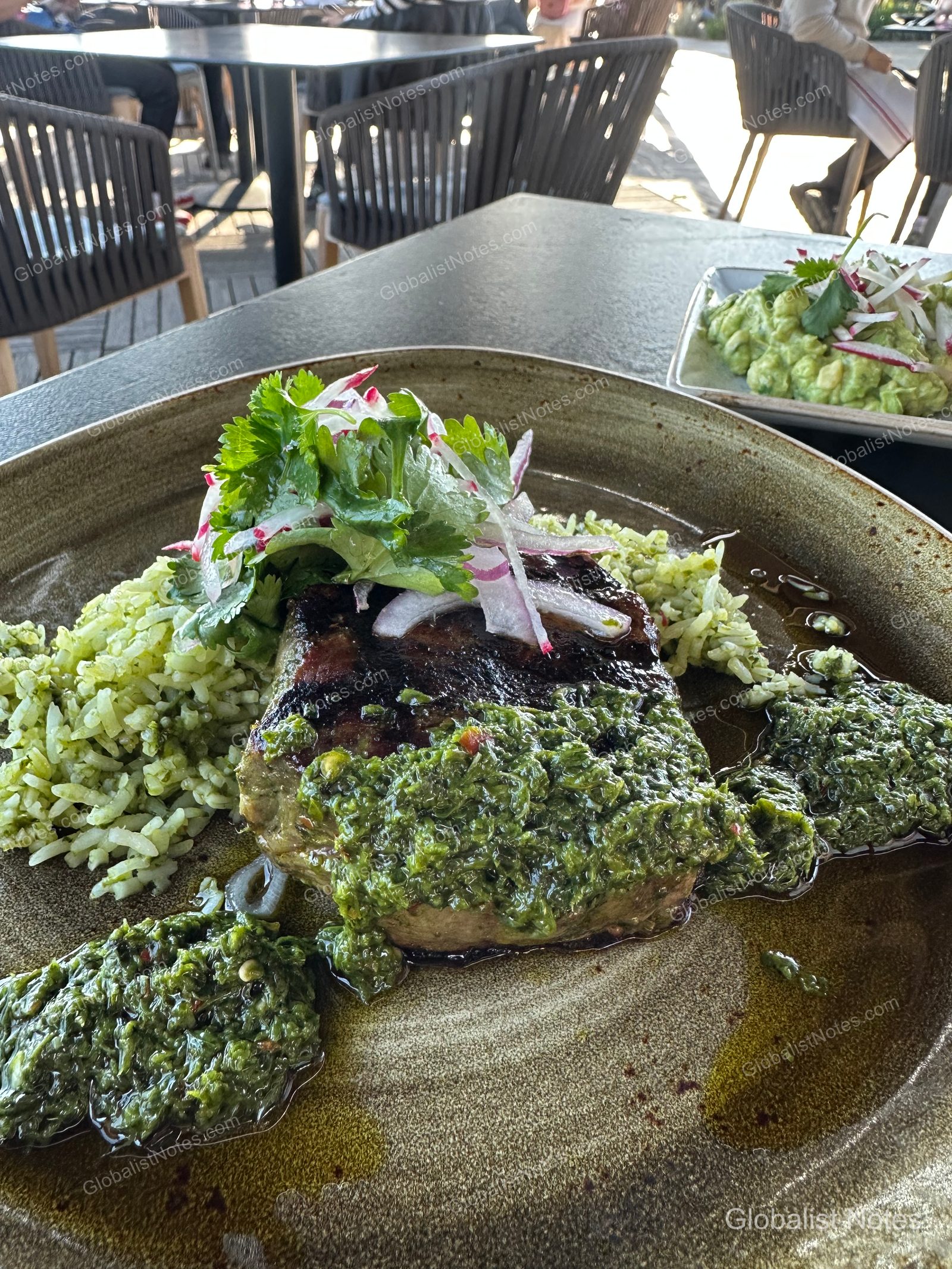A lunch plate on an outdoor table at Alila Ventana Big Sur, featuring grilled steak topped with chimichurri sauce, served with green herb rice, cilantro, and sliced red onion. Outdoor terrace furniture is visible in the blurred background.