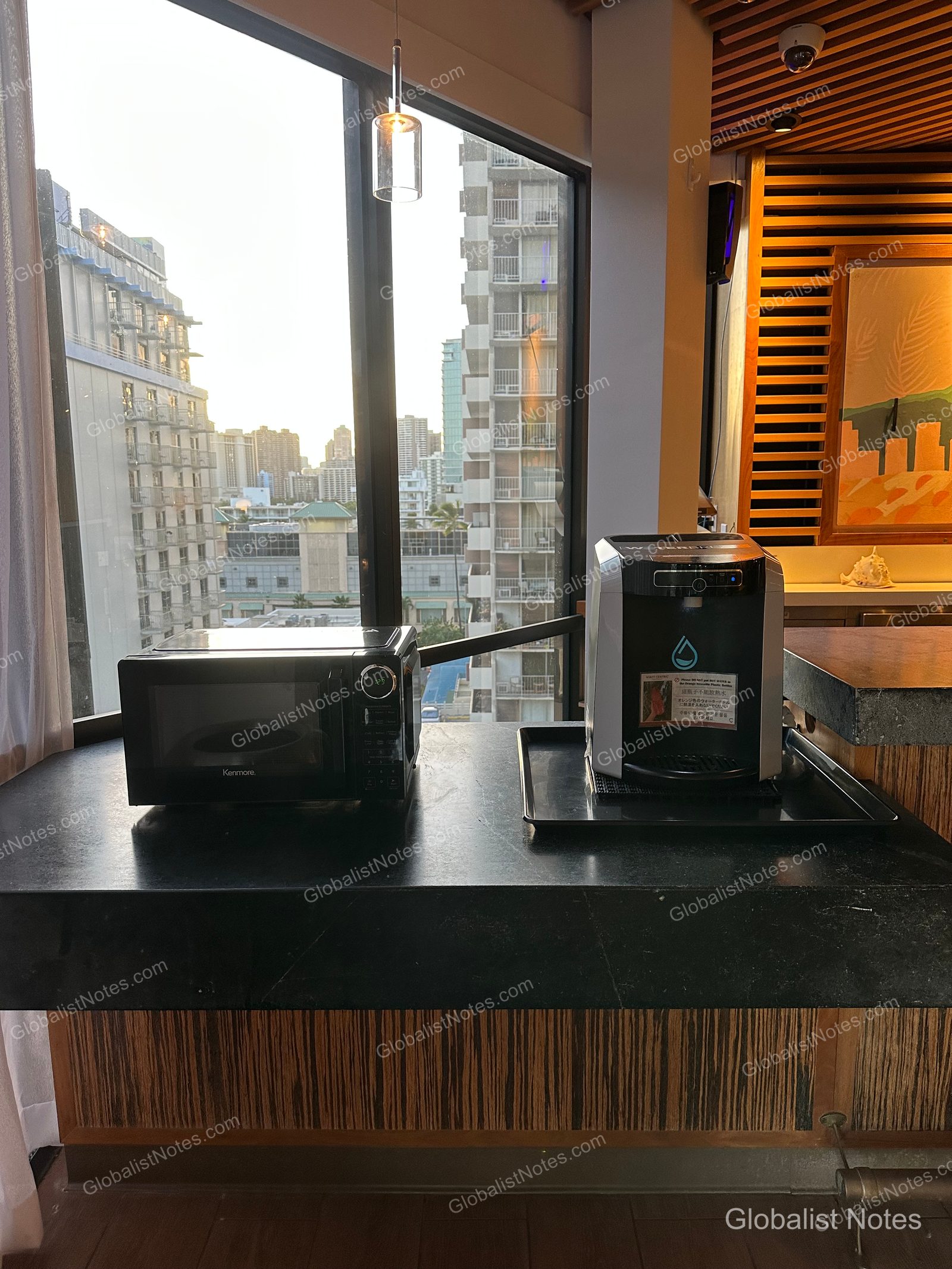 A microwave and water dispenser placed on a black counter in the common area of Hyatt Centric Waikiki Beach. High-rise buildings in downtown Honolulu are visible through the window, and the appliances are neatly arranged under indoor lighting.