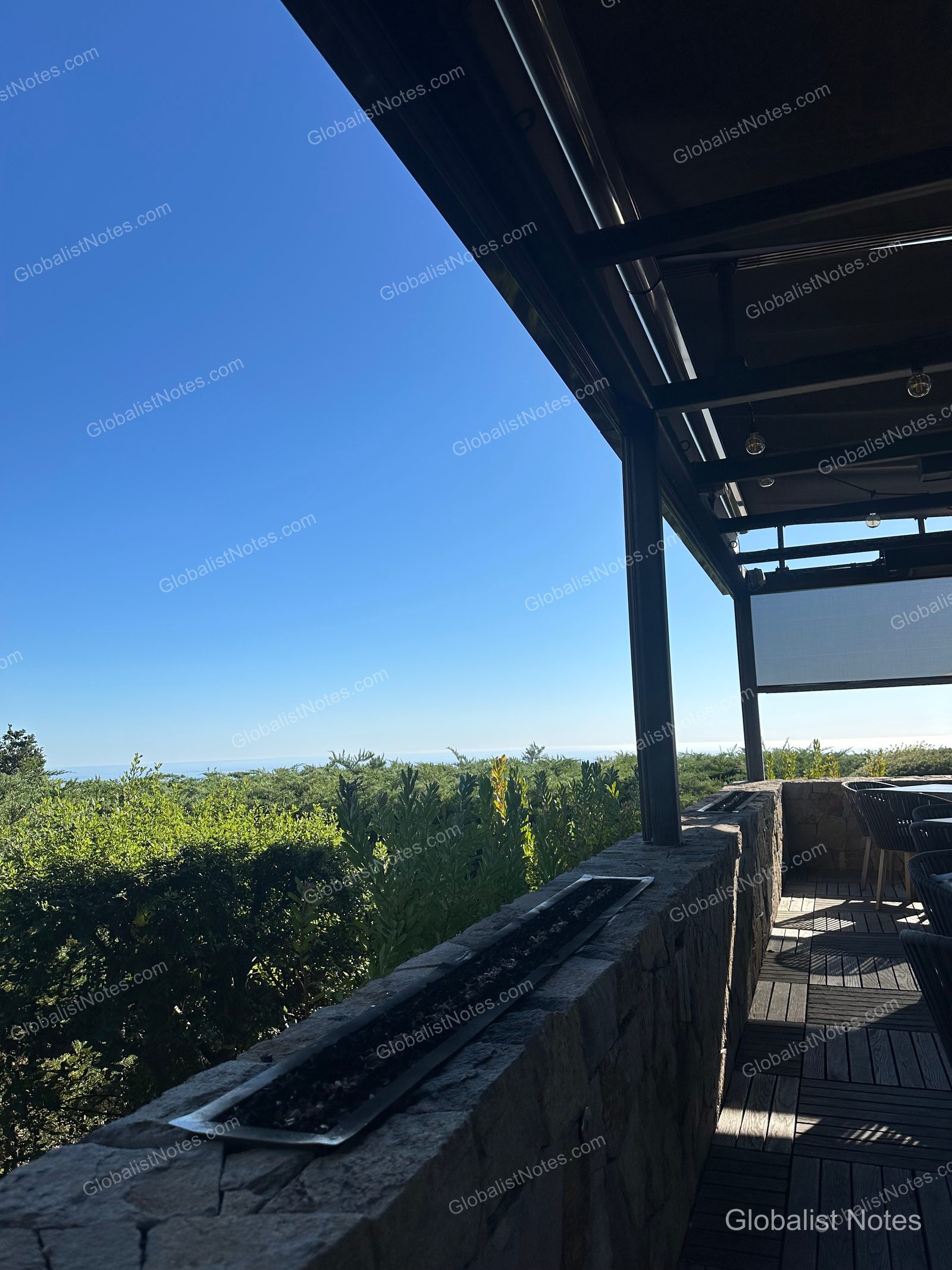 View from the outdoor terrace at Alila Ventana Big Sur, featuring a long fire pit on a stone railing, with the blue sky, dense forest, and the horizon visible in the distance.