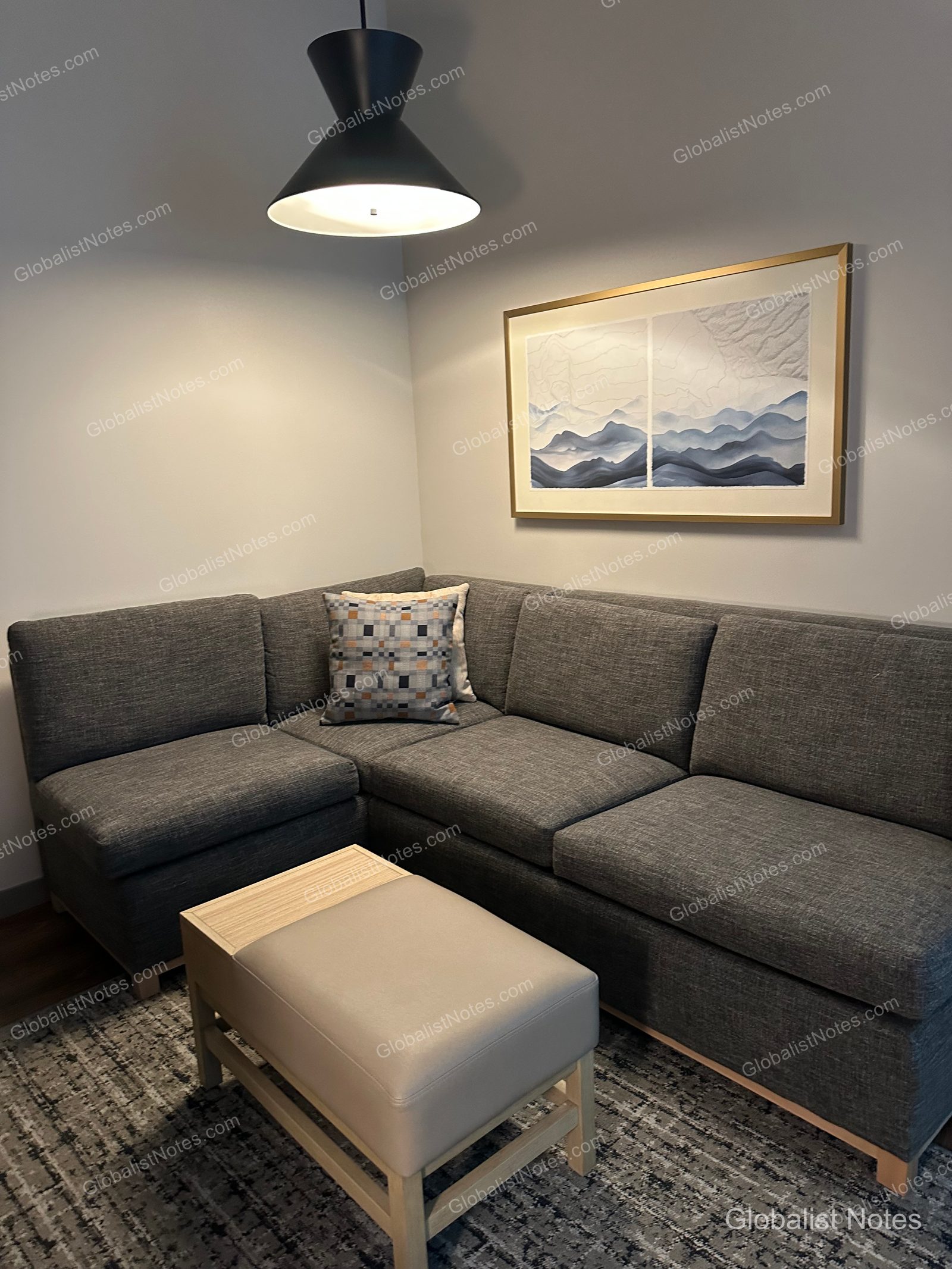 Hyatt Place Bozeman Yellowstone Airportのジュニアスイートのコーナーエリア。グレーのL字型ソファとベージュのオットマンが置かれ、上部にはモダンなペンダントライト、壁面には山脈をモチーフにしたアートが飾られています。