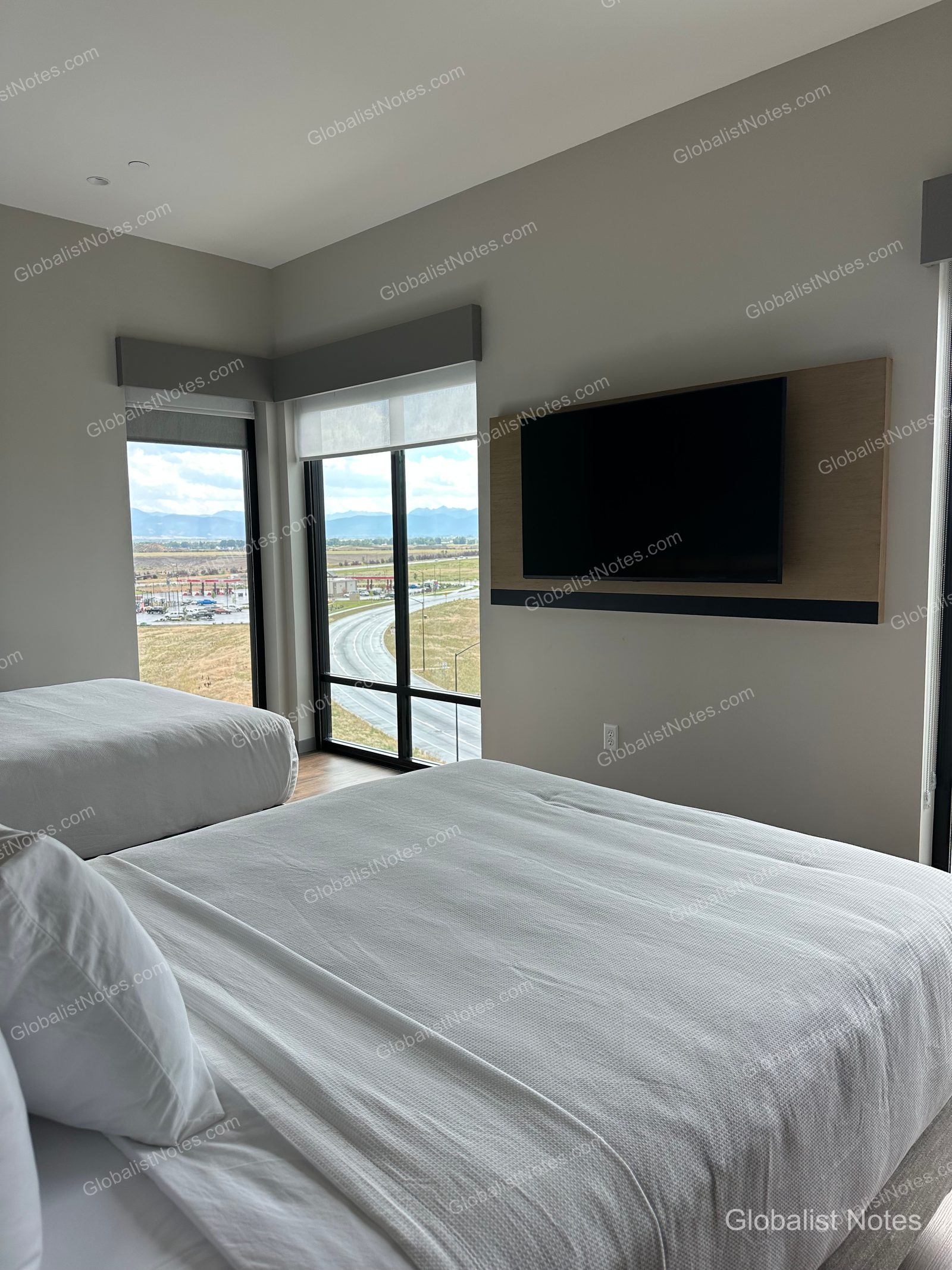Hyatt Place Bozeman Yellowstone Airportのジュニアスイートルーム。白い寝具が整えられたベッド2台が並び、壁面には大型テレビが設置されています。コーナーの大きな窓からは、ベルグレードの街並みと遠くの山々が見渡せます。