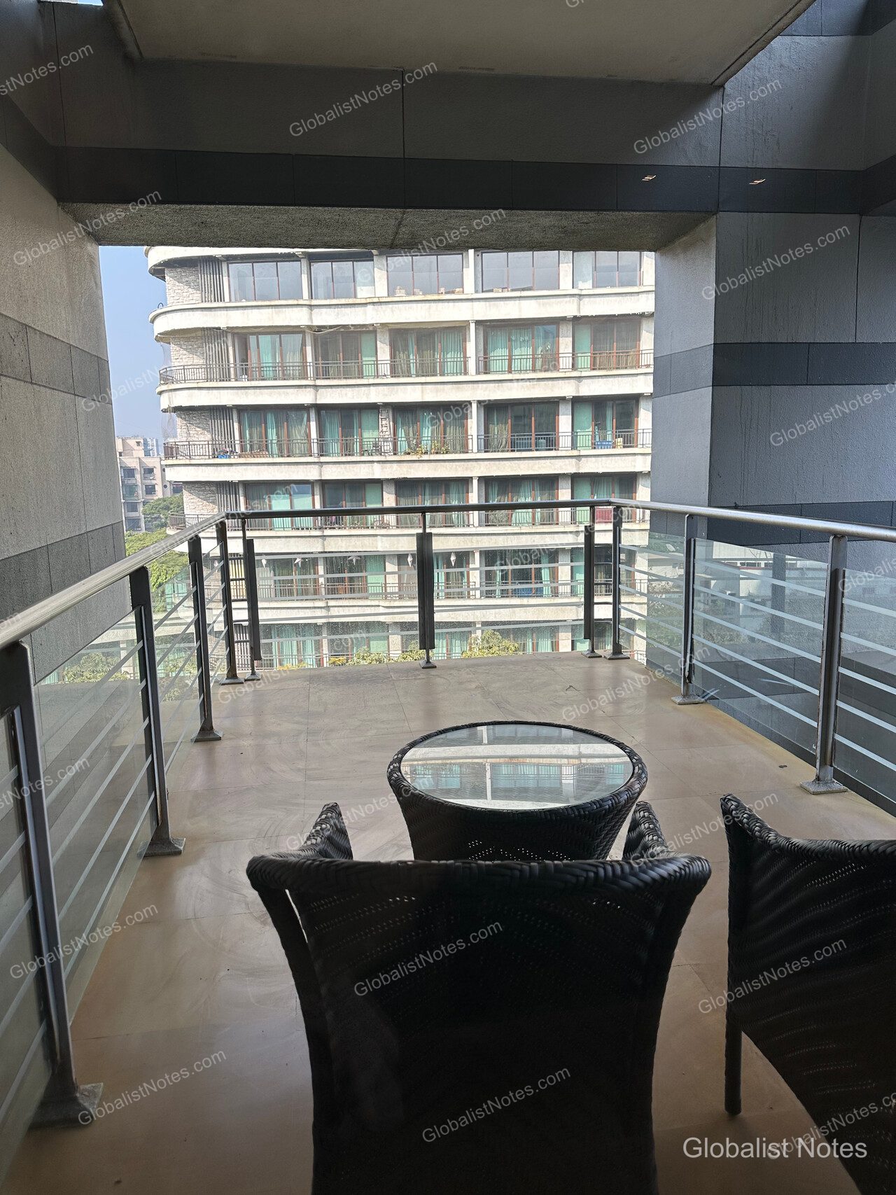 The spacious outdoor terrace of the Veranda Suite King at Grand Hyatt Mumbai Hotel & Residences, featuring two dark brown rattan chairs and a glass-topped round table, with views of neighboring hotel buildings.